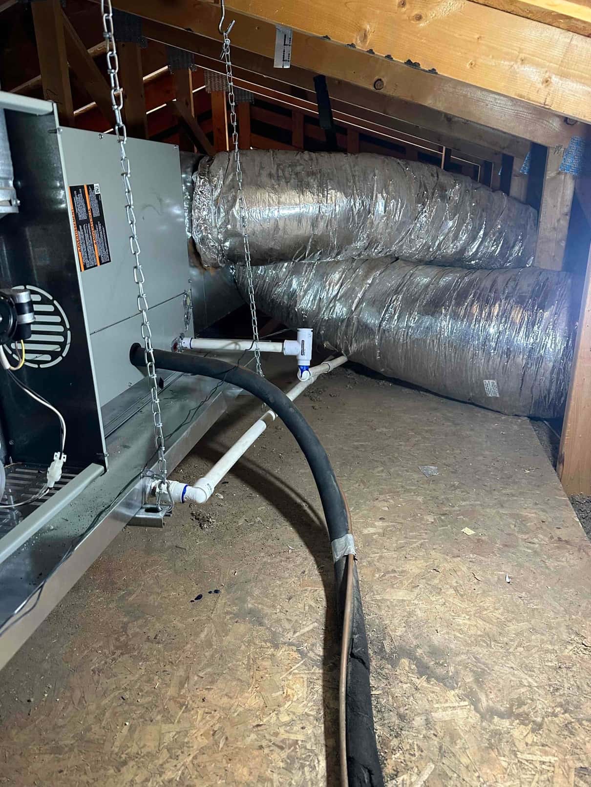 Air handler installed in attic with insulated flexible duct connections, suspended by chains, condensate drain with cleanout, and insulated refrigerant line set.