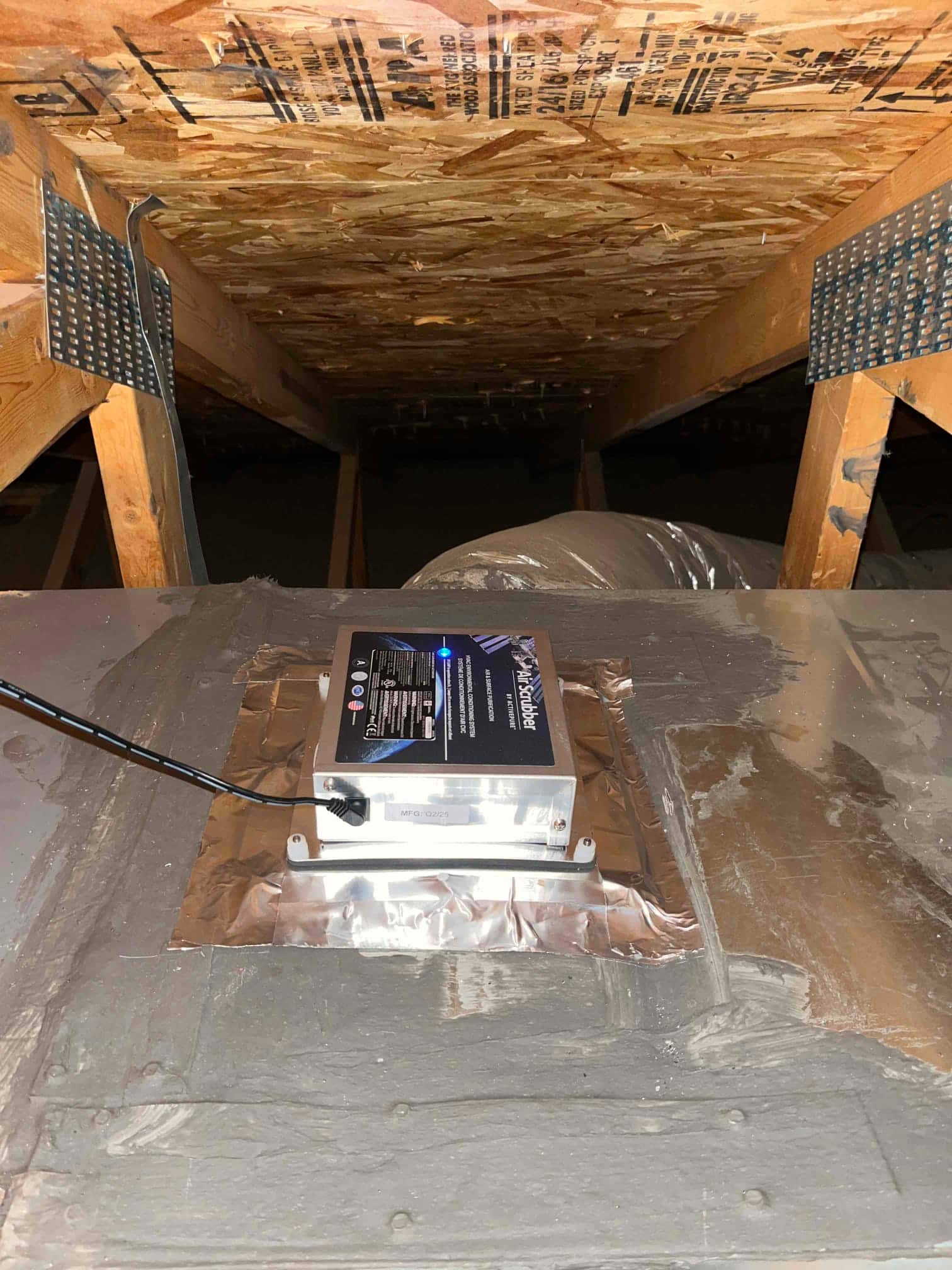 Air scrubber unit installed on the duct surface in the attic, sealed with foil tape around the mounting plate.