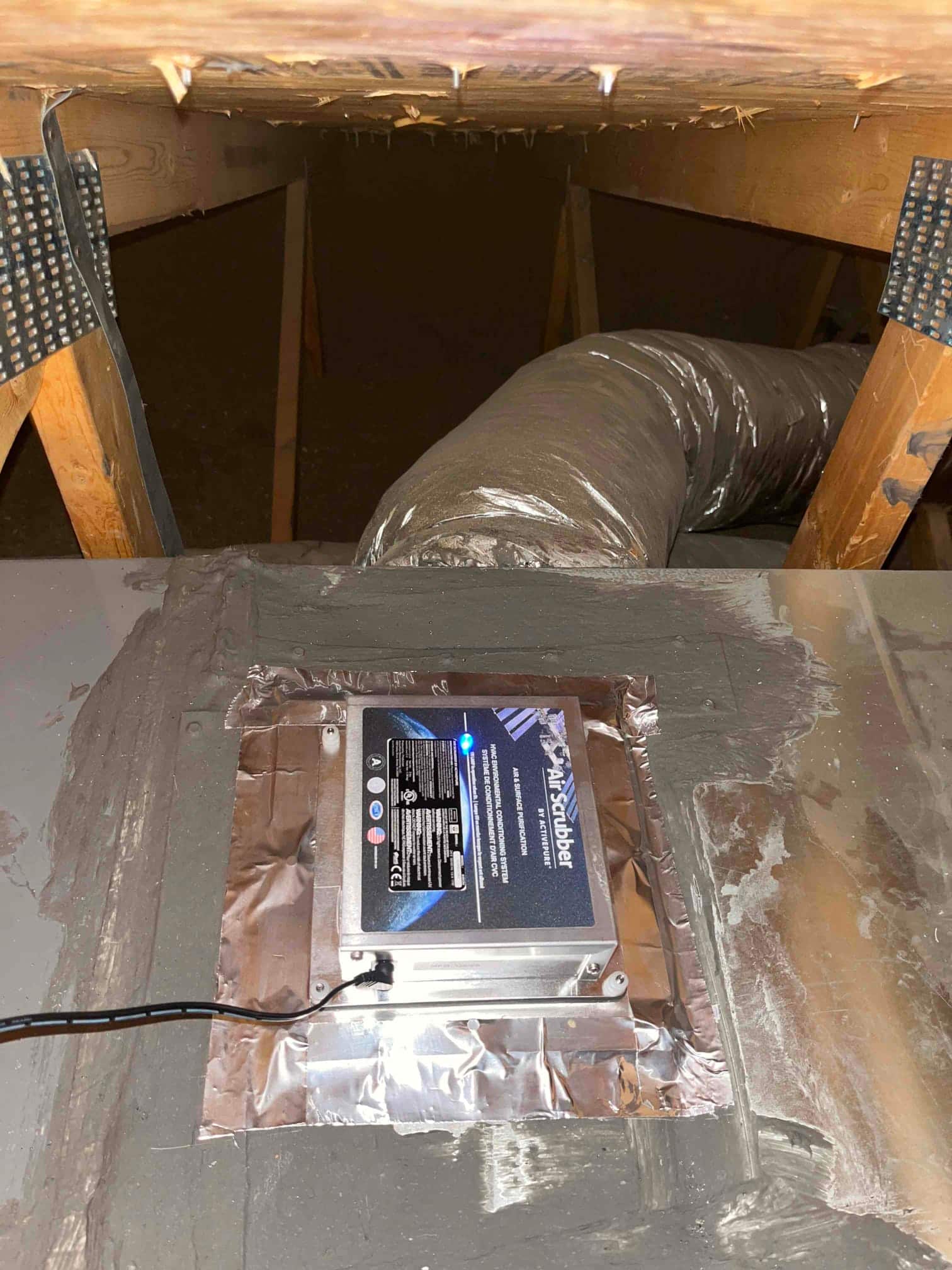 Air scrubber unit installed on ductwork in the attic, sealed with foil tape, blue status light illuminated.