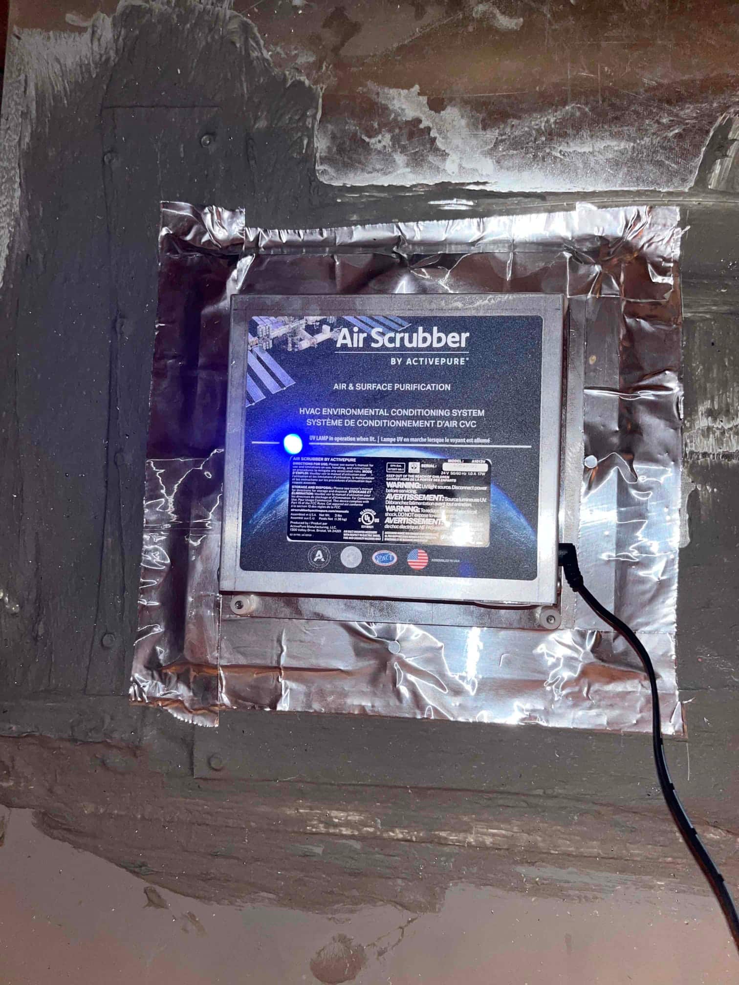 Air scrubber unit mounted on ductwork with foil tape sealing edges and power indicator light illuminated.