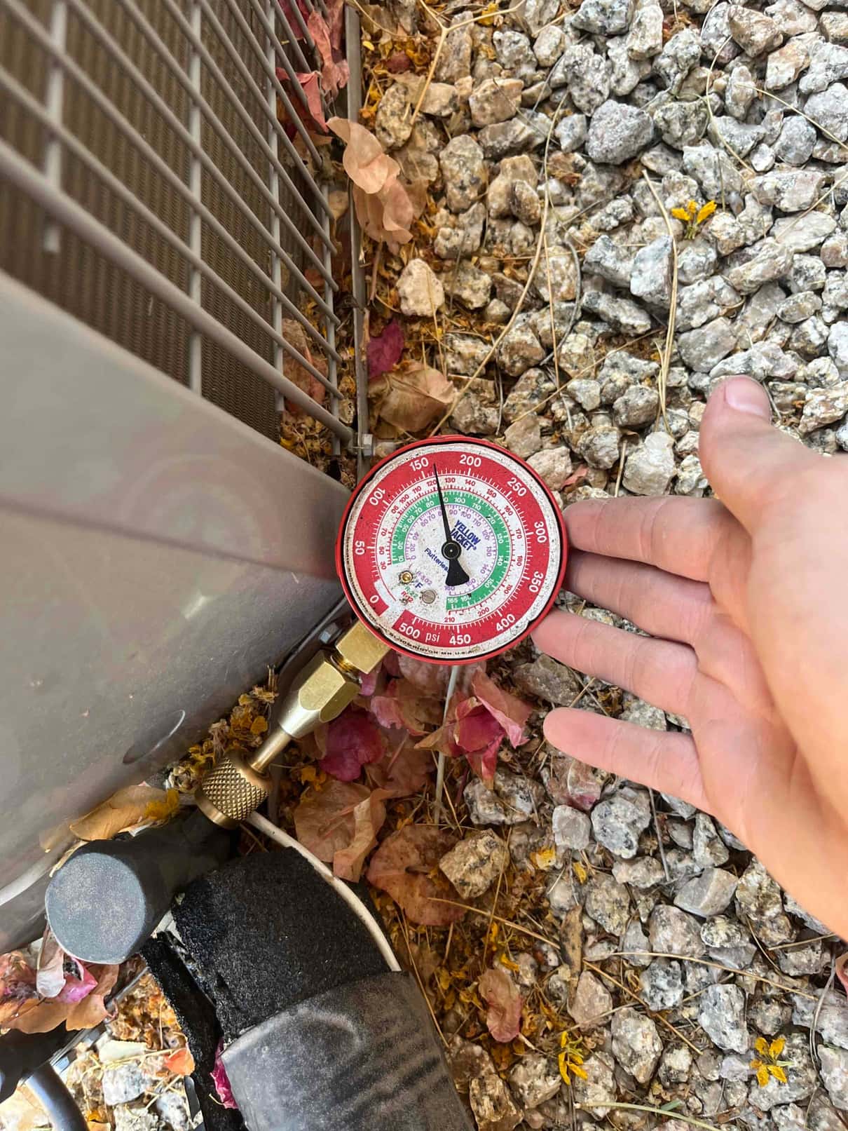 Analog refrigerant gauge connected to the service port on an outdoor condenser unit during diagnostic pressure check.