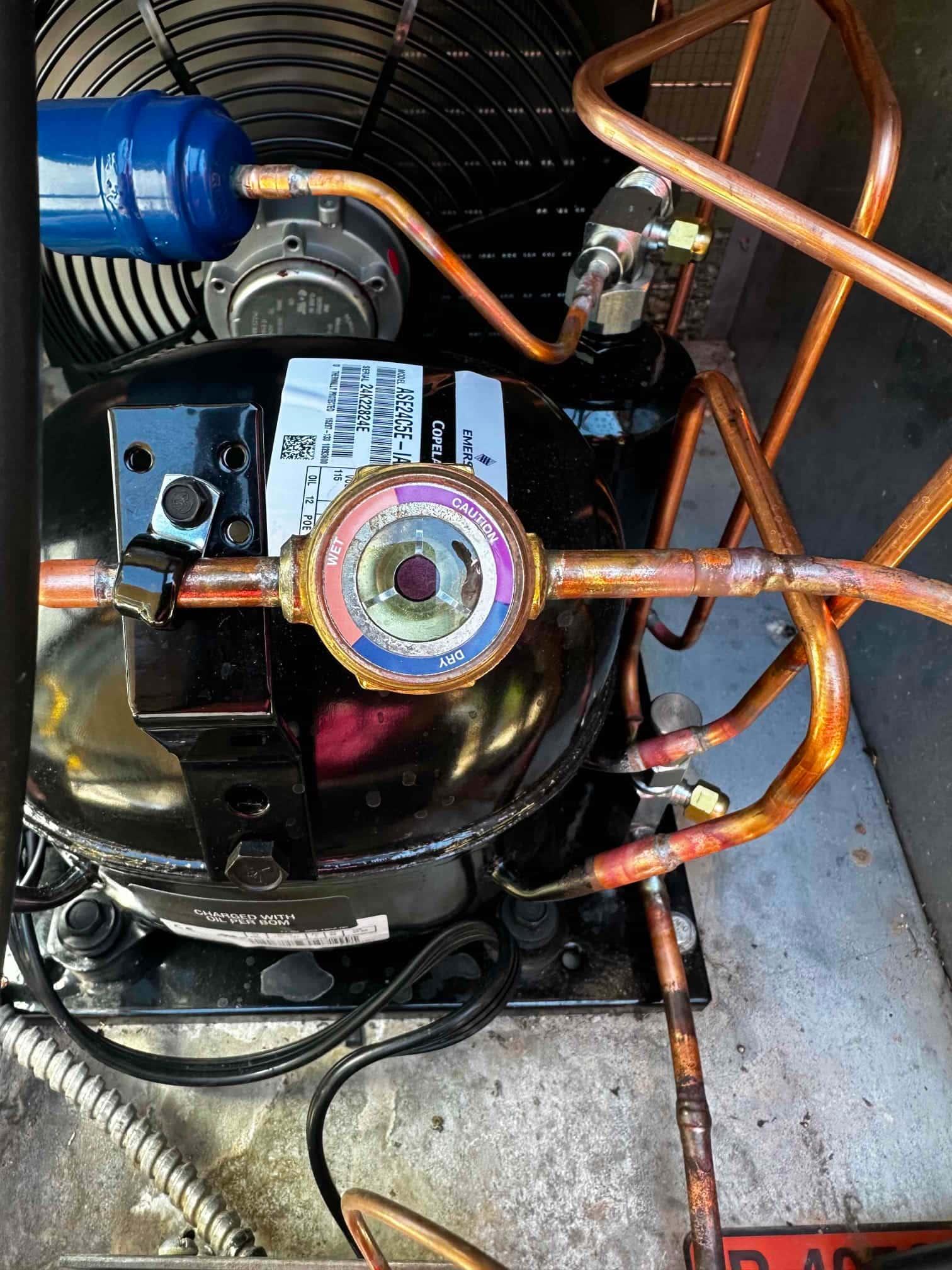Close-up of a new condenser’s internal components, showing copper refrigerant lines, filter-drier, sight glass, and compressor housing.