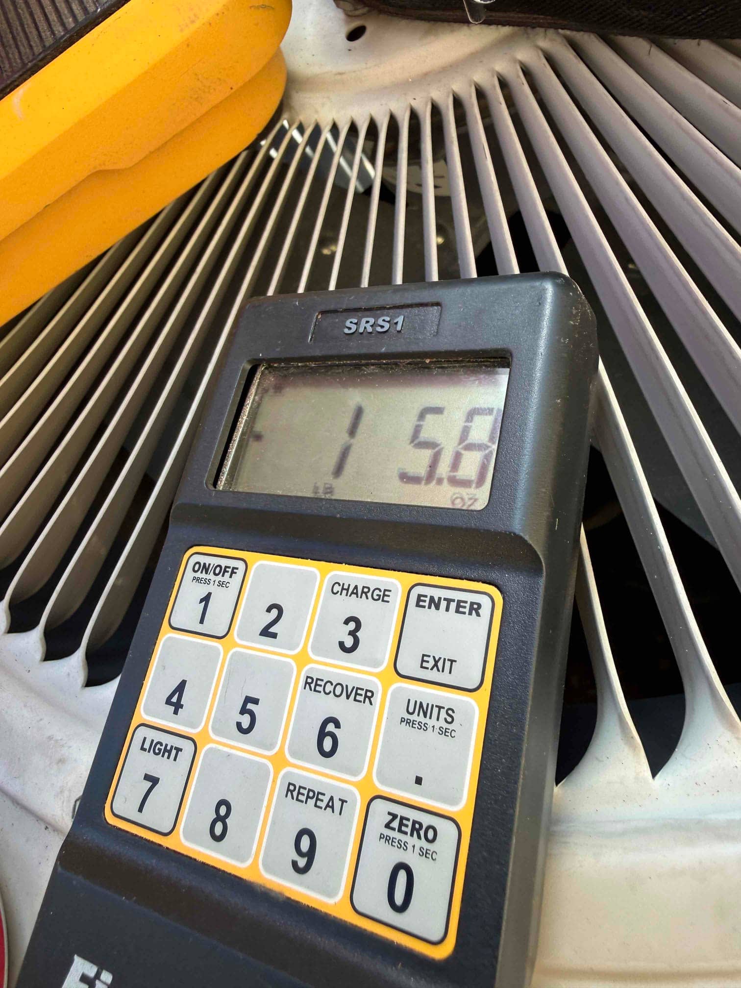 Digital refrigerant scale on the outdoor unit fan grille displays a reading while charging controls are visible on the keypad.