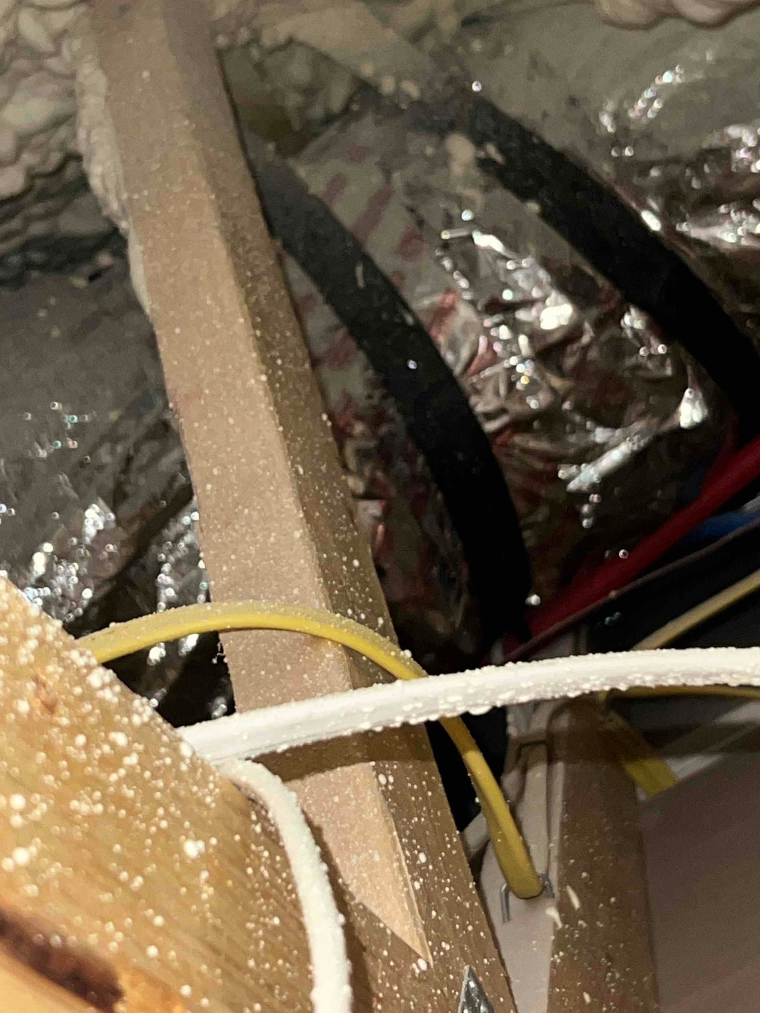 Foil-insulated flexible duct runs through attic space with black straps and nearby electrical wiring visible.