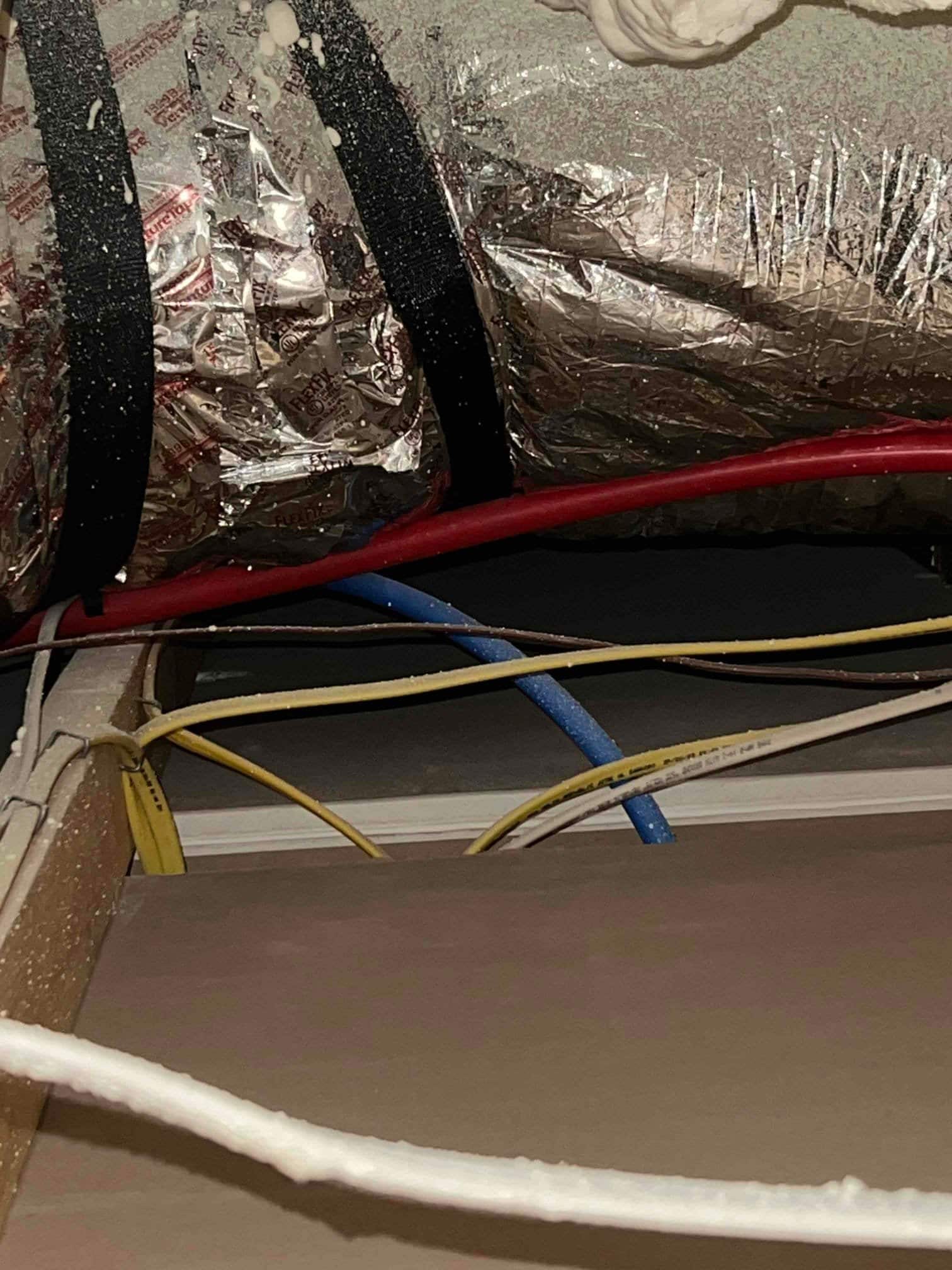 Foil-insulated round duct with black straps and nearby electrical wiring visible in the attic space.