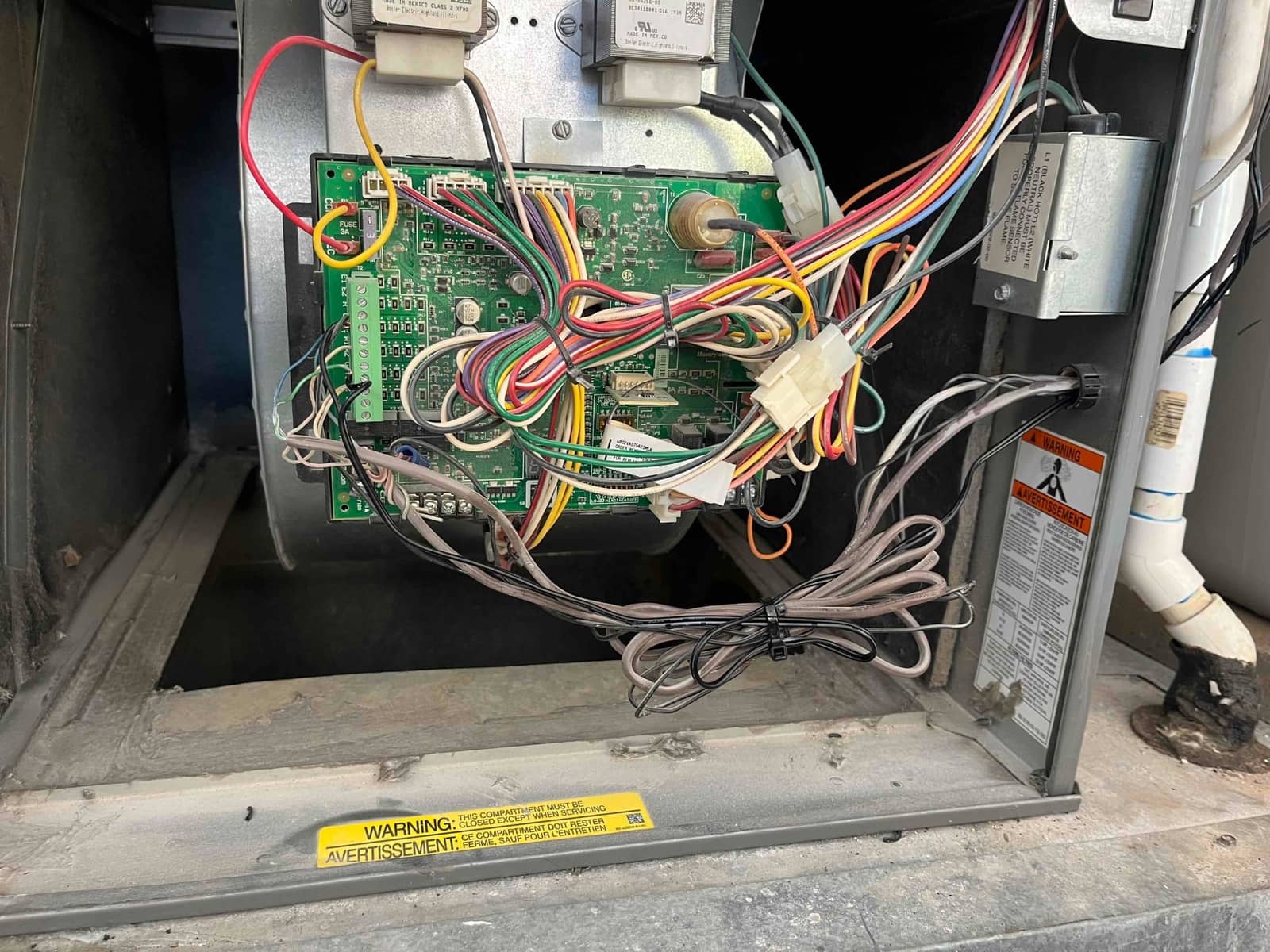 Furnace control board exposed with multi-colored wiring harnesses connected; blower housing, transformer, and safety labels visible inside the air handler.