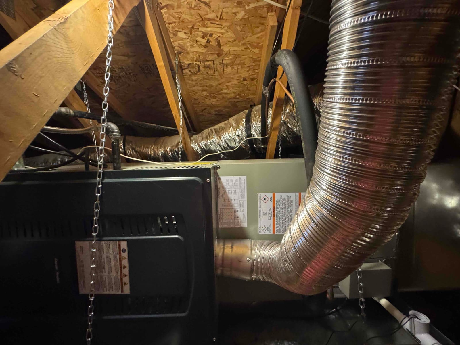 Horizontal air handler suspended in attic with insulated flex duct connections, sheet-metal transition, condensate drain, and hanging support chains.