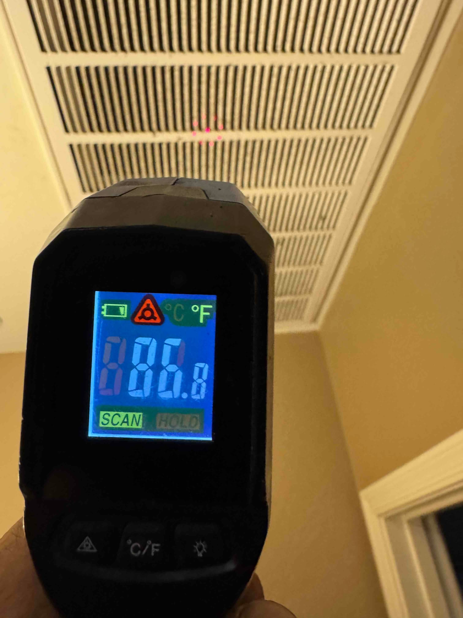 Infrared thermometer aimed at a return grille, display illuminated but temperature digits not readable for documentation.