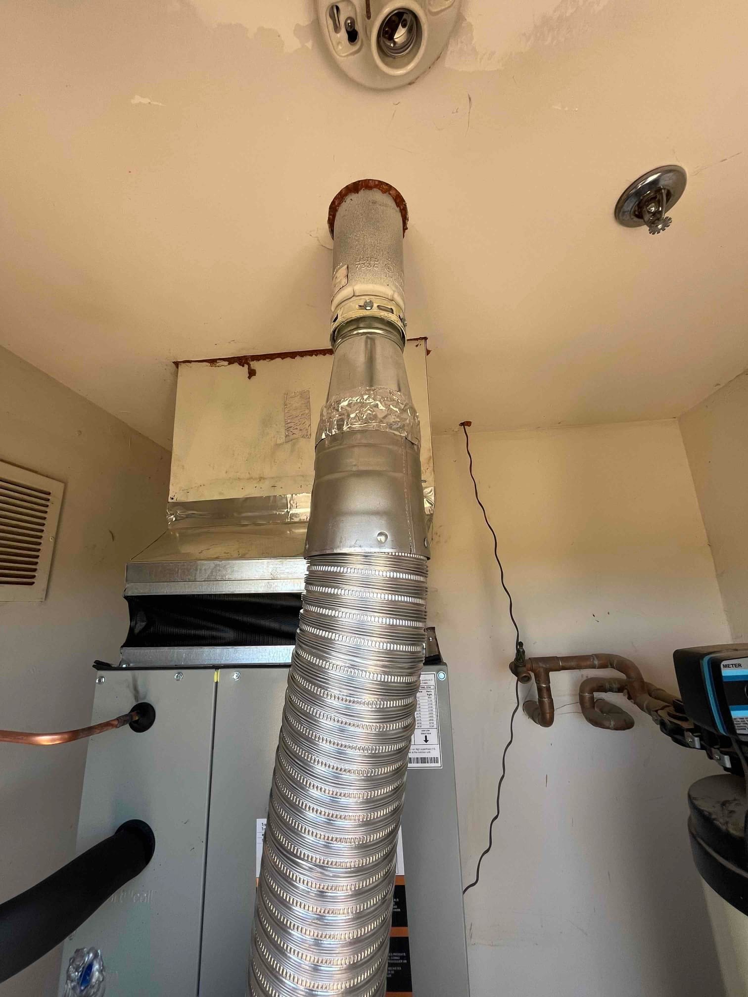 New gas furnace installed with vent connector and flue pipe routed to ceiling, connected to duct plenum and refrigerant lines.
