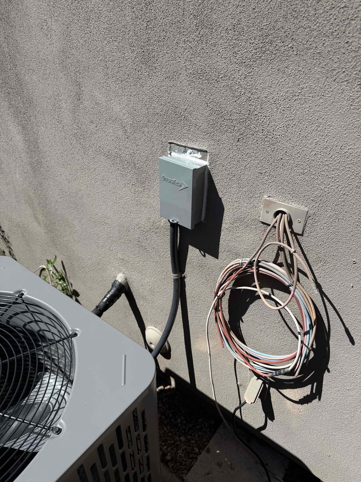 Outdoor condenser next to newly mounted disconnect and sealed wall penetrations, with thermostat control wiring coiled beside the unit.