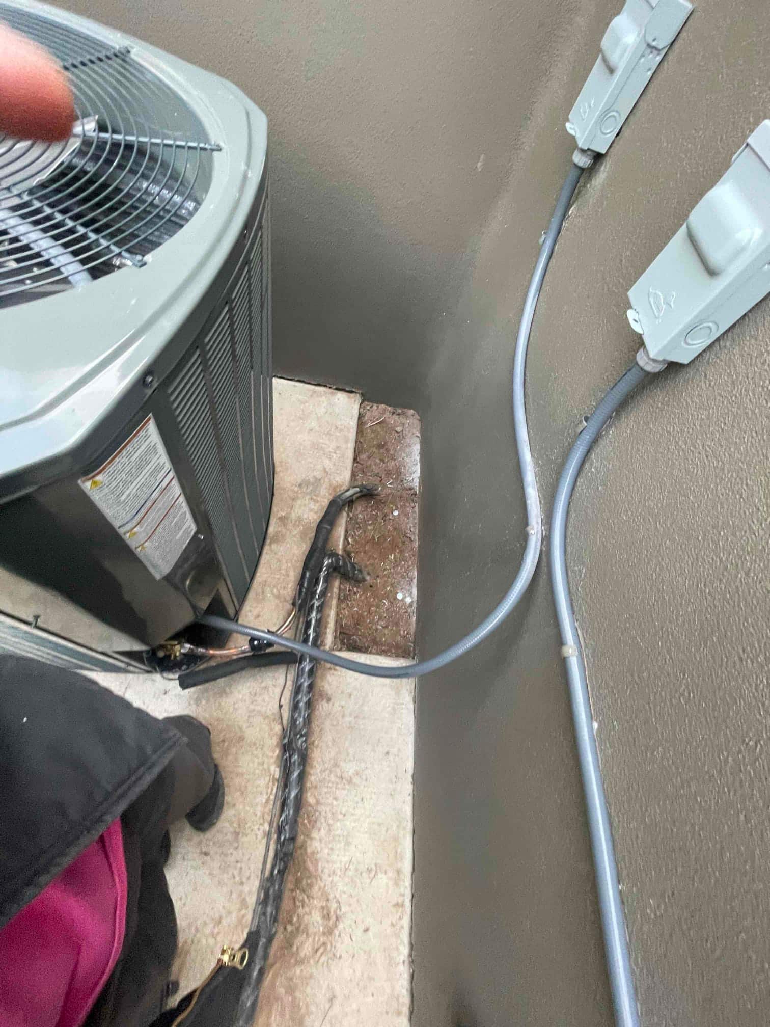 Outdoor condenser set beside wall with insulated refrigerant lines and two electrical disconnect boxes installed in conduit.