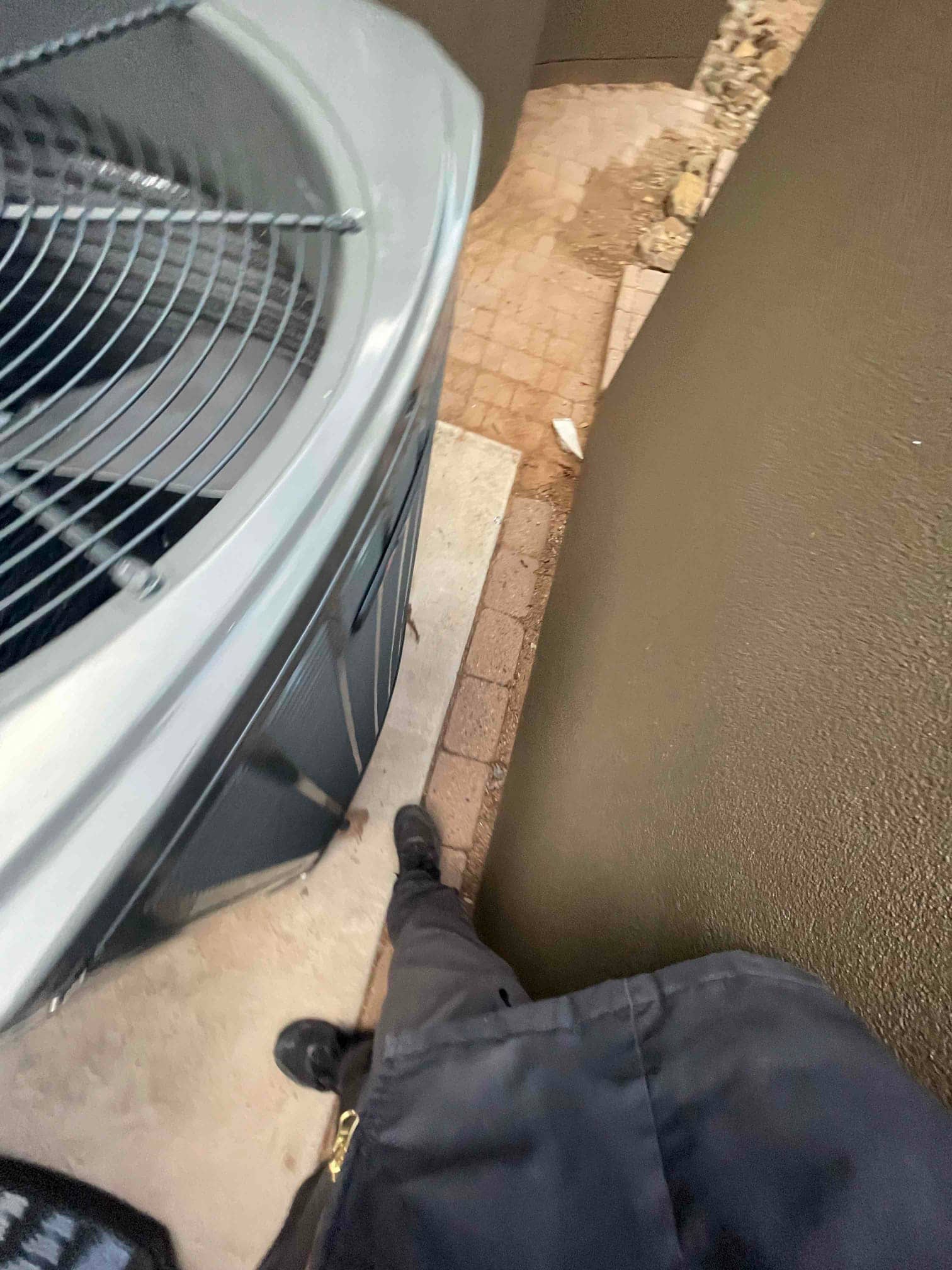 Outdoor condenser set on a concrete pad in a narrow side yard, with protective top grille visible beside a technician’s legs.