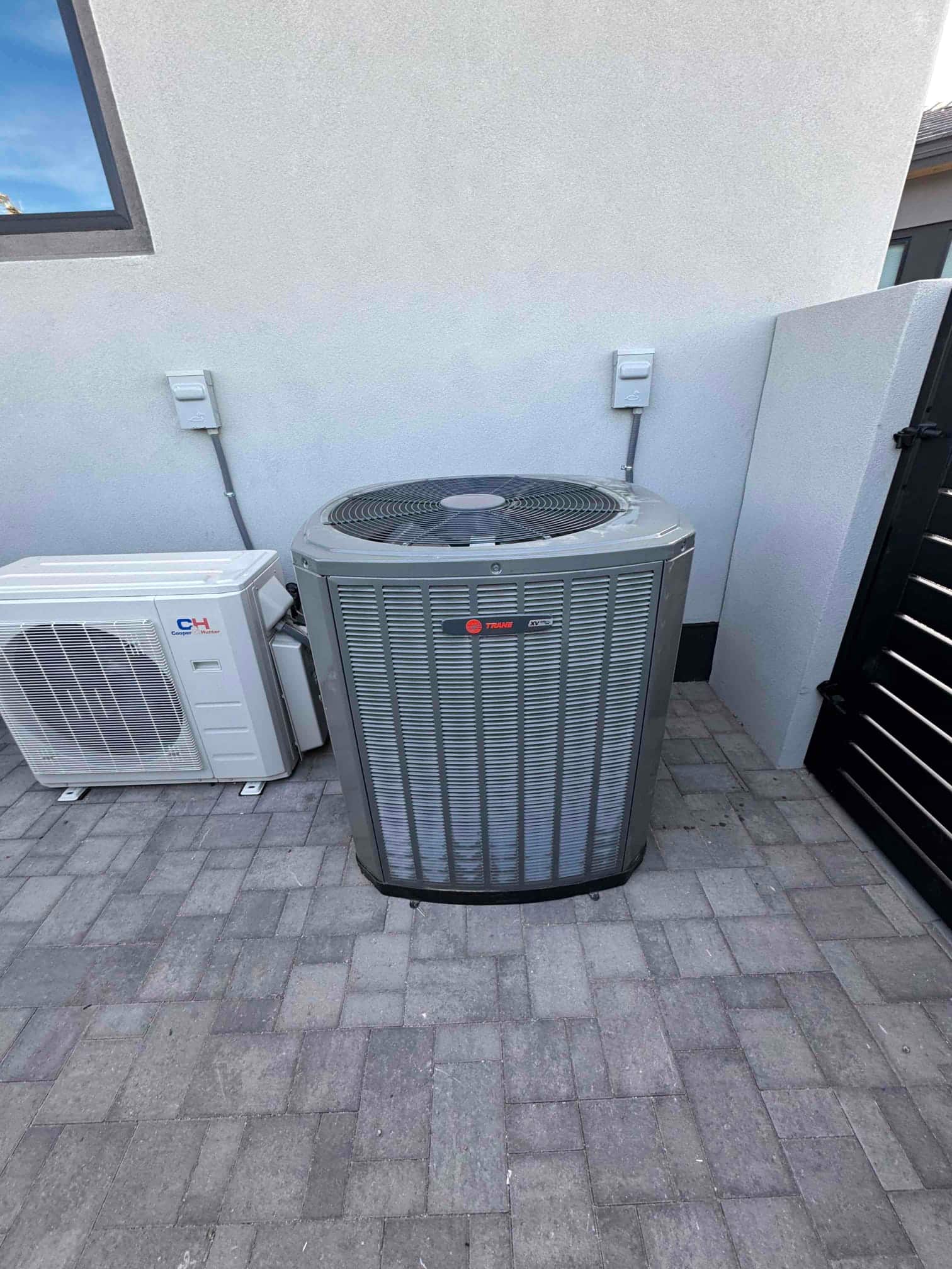 Outdoor condenser set on a paved pad with adjacent mini-split unit and installed electrical disconnects on the wall.