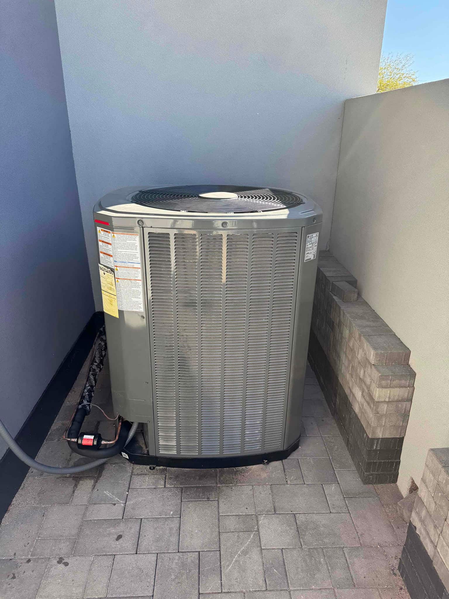 Outdoor condenser set on a paved surface with insulated refrigerant lines and electrical whip connected along the wall.