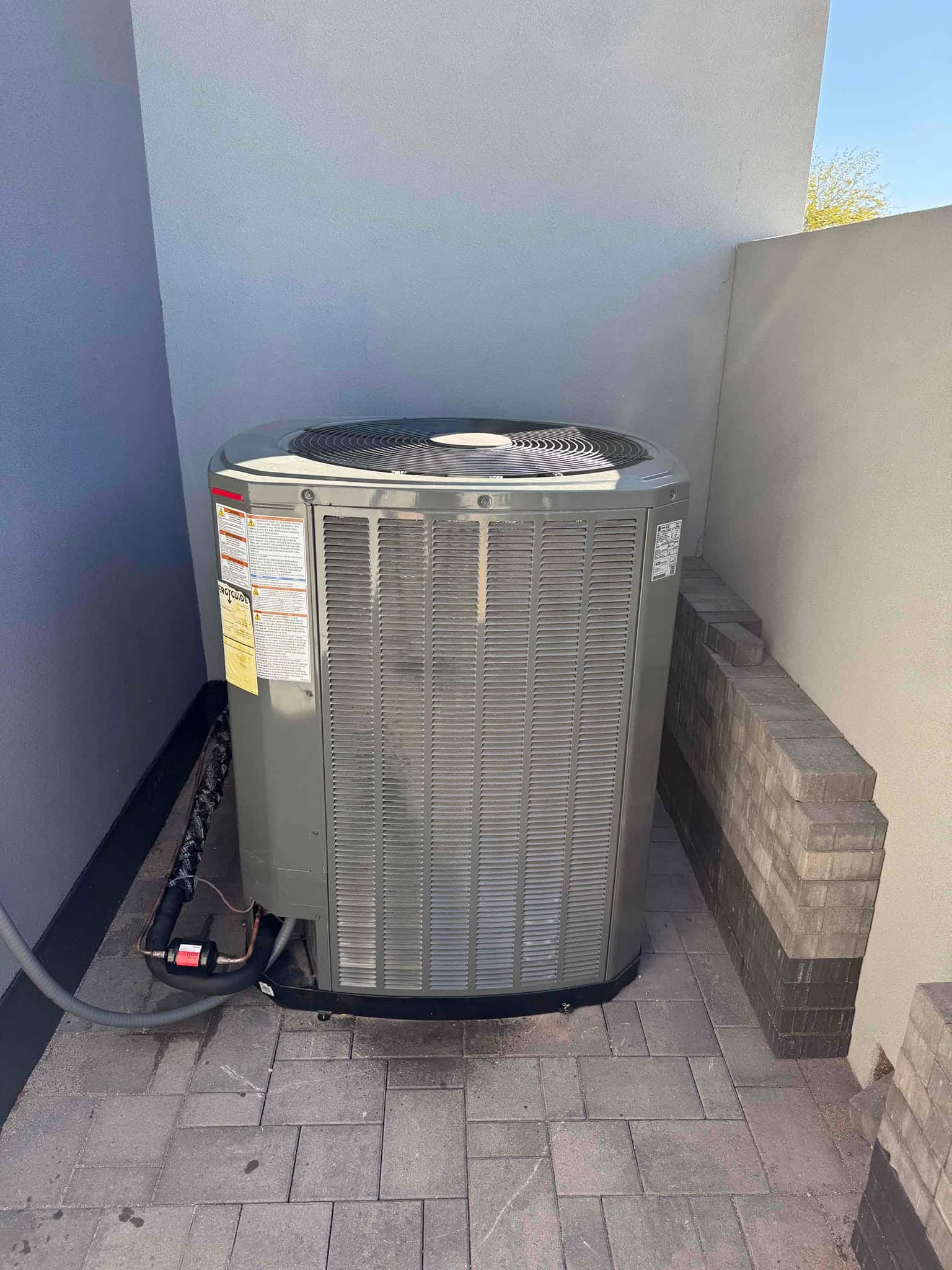 Outdoor condensing unit installed on a paved pad with insulated refrigerant lines and electrical whip routed to the cabinet.