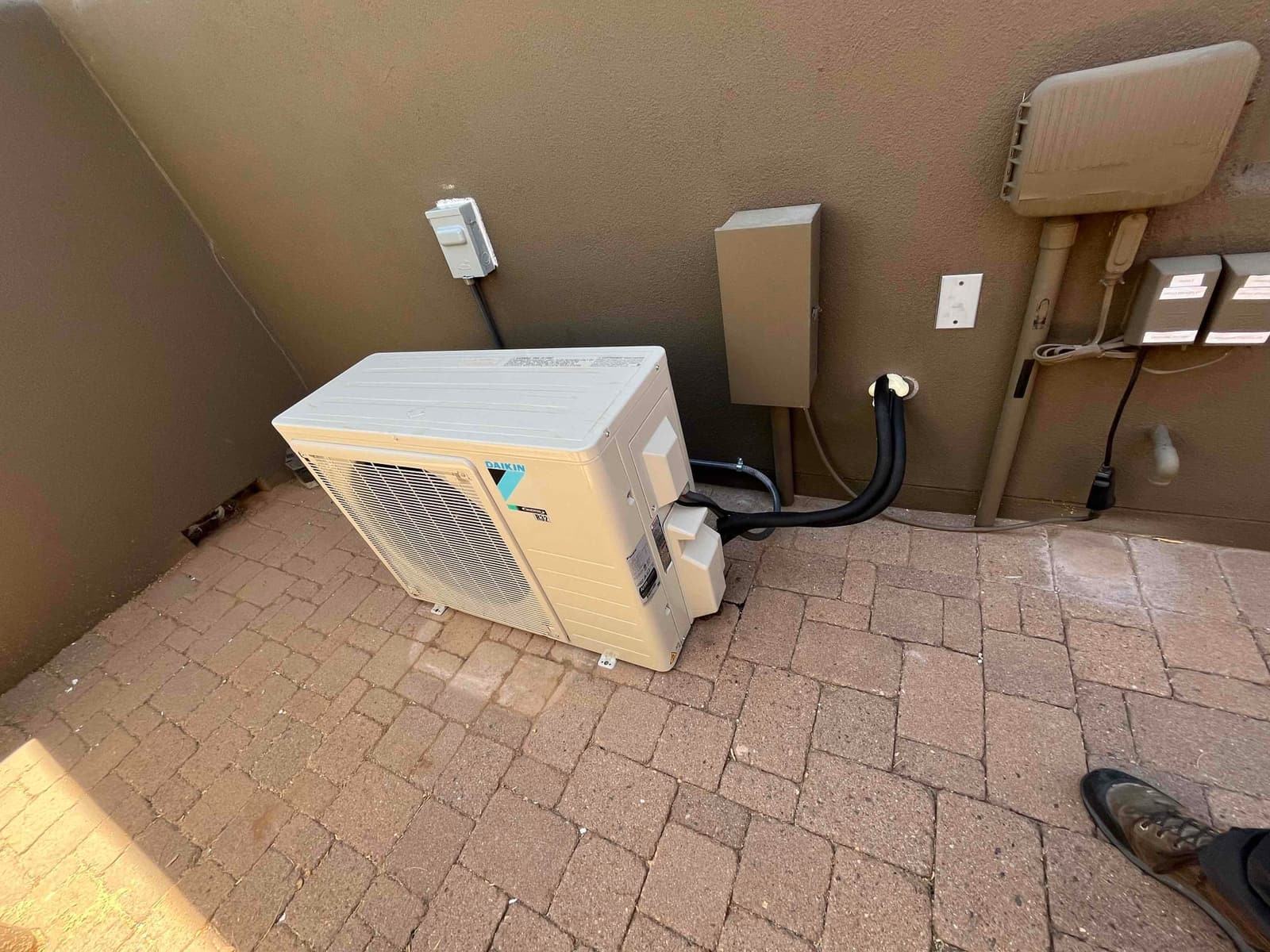 Outdoor mini-split condenser set on paver surface with insulated line set routed through wall and electrical disconnect mounted nearby.