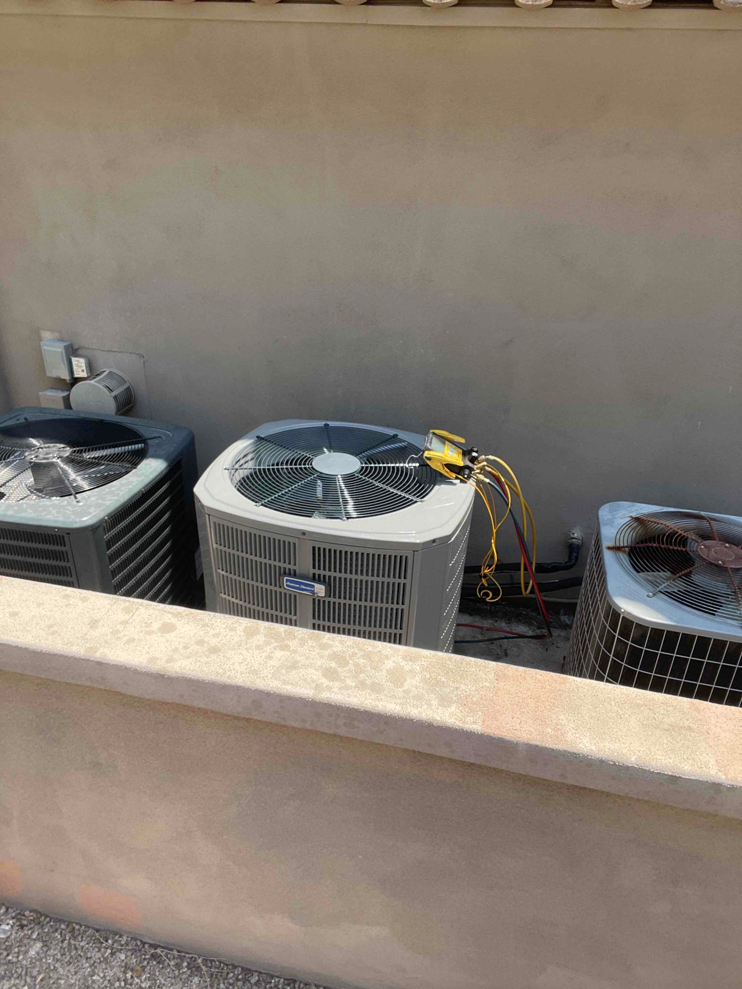 Row of outdoor condensers behind a low wall, with manifold hoses connected to the middle unit during installation.