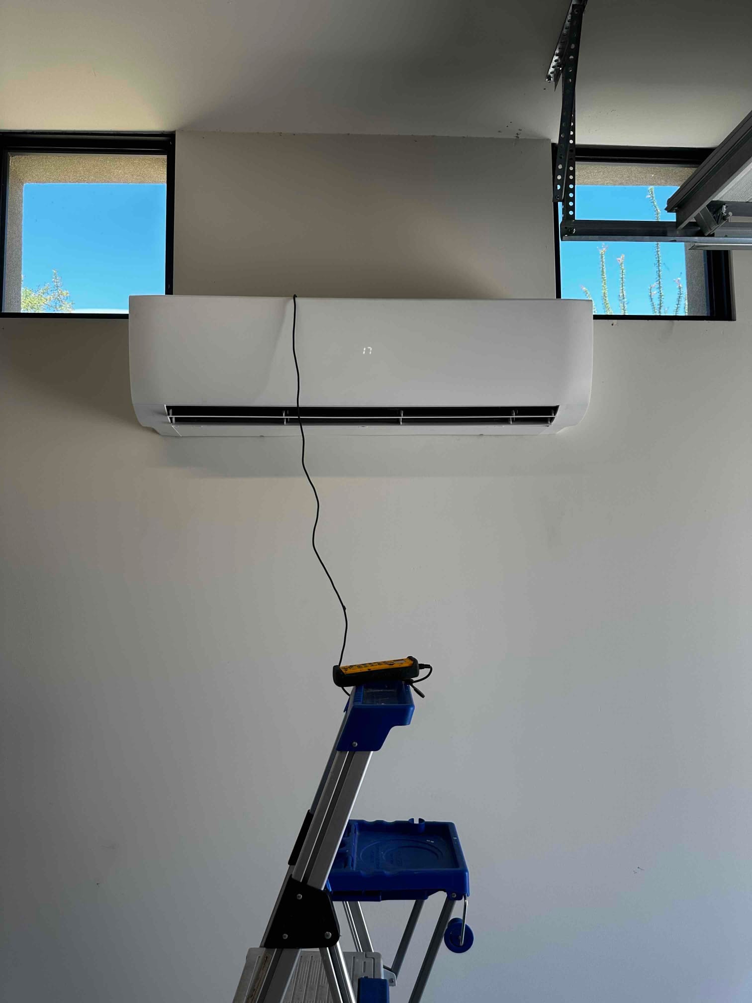 Wall-mounted mini-split indoor unit newly installed, with ladder and tool beneath for final setup and testing.