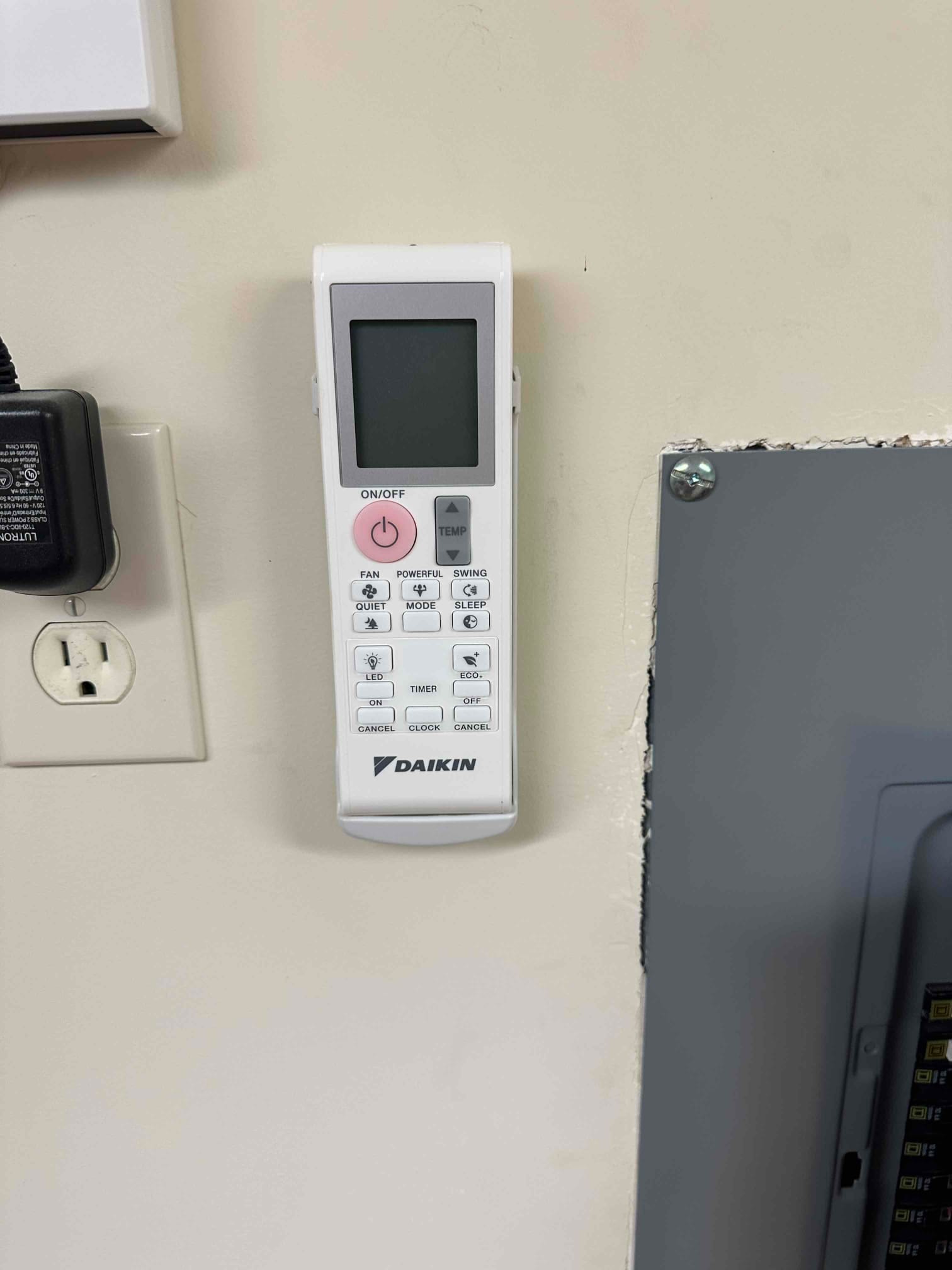 Wall-mounted mini-split remote control in its holder next to an electrical panel and outlet after installation.