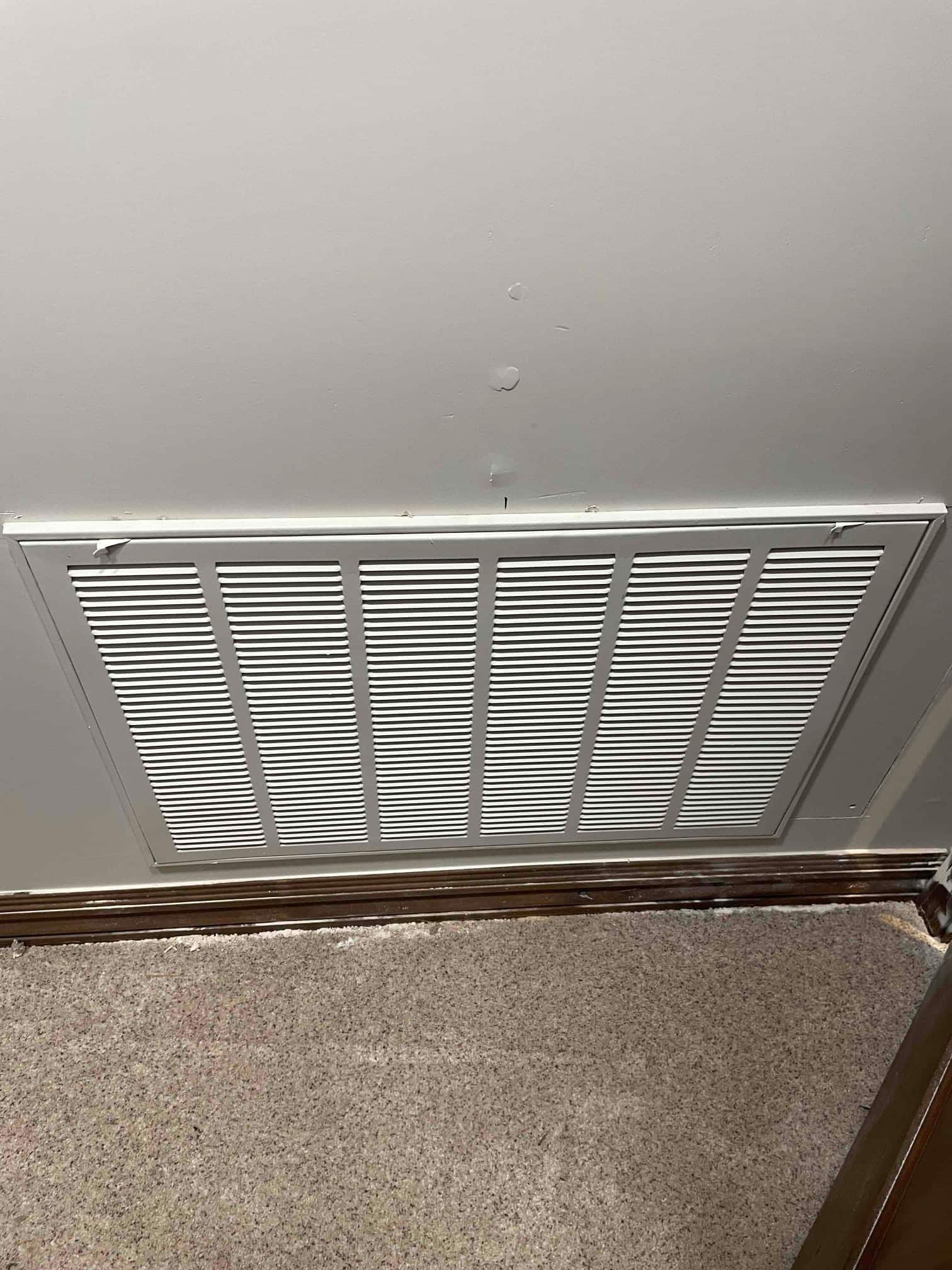 Wall-mounted return air grille installed above baseboard with surrounding trim and carpeted floor visible.