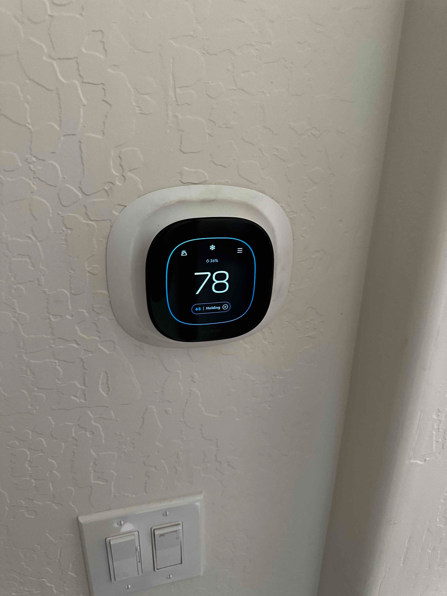 Wall-mounted smart thermostat installed and operating, displaying a setpoint screen with illuminated interface above nearby light switches.
