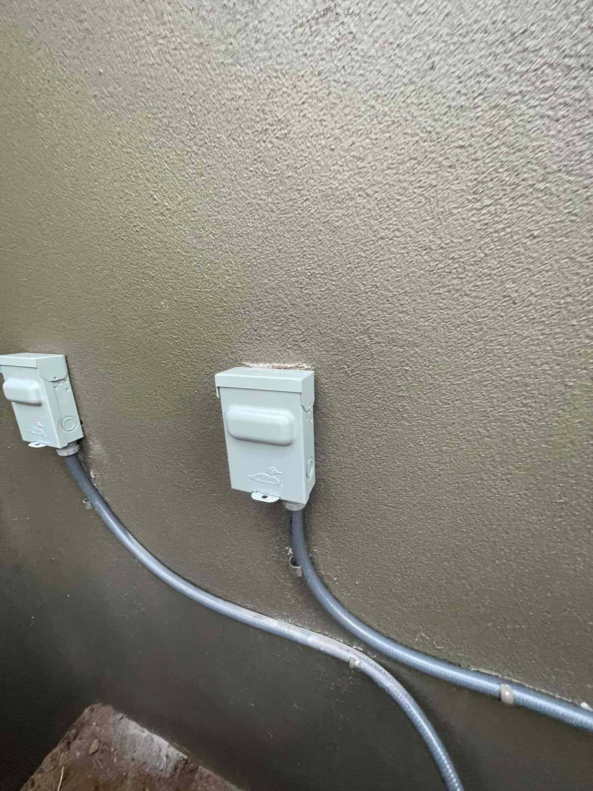 Weatherproof electrical disconnect boxes mounted on exterior wall with flexible conduit secured along the surface for HVAC equipment.