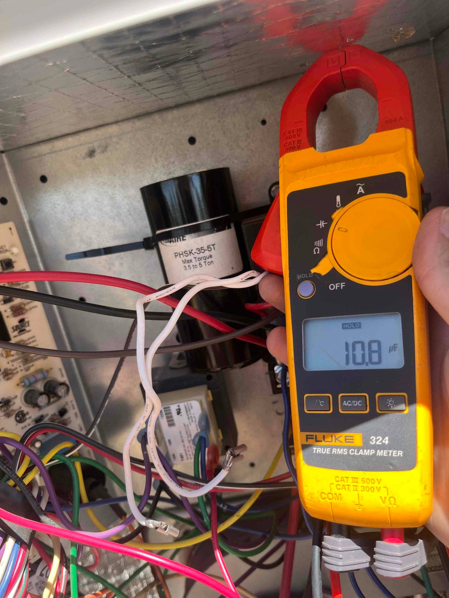 Clamp meter displays 10.8 µF while measuring a capacitor inside the outdoor condenser control compartment with wiring exposed.