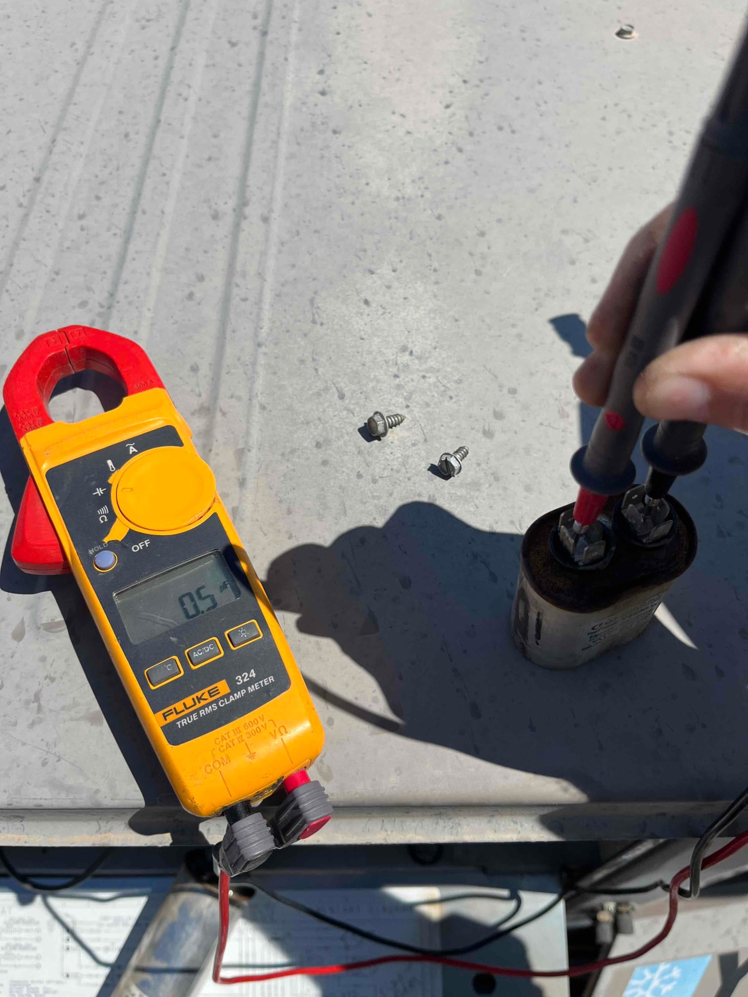 Clamp multimeter displays 0.51 A while probes contact terminals on the removed dual‑run capacitor during testing.