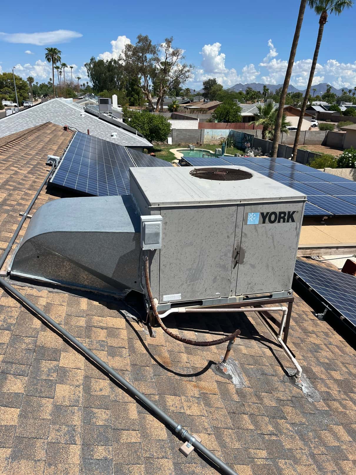 Package air conditioning unit on a shingle roof with connected duct transition and electrical disconnect, surrounded by rooftop solar panels.