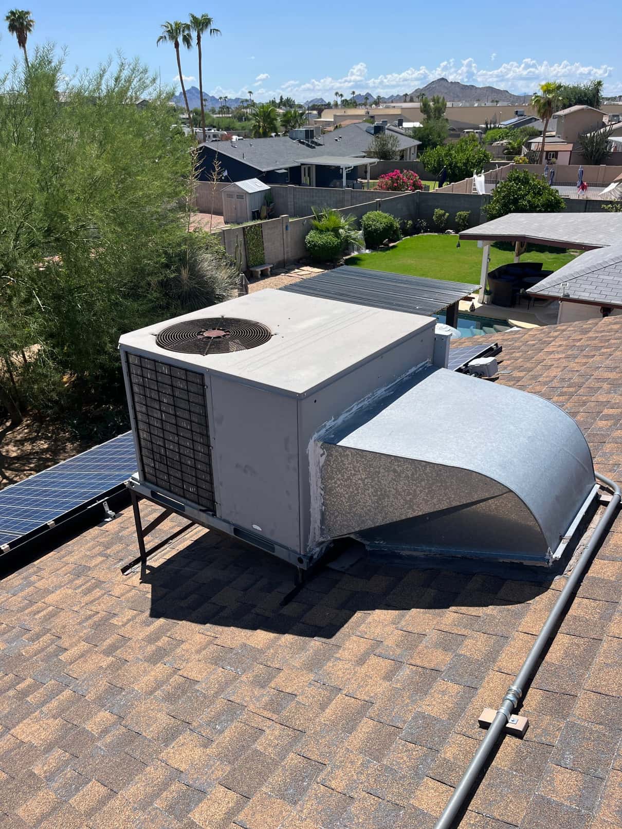 Rooftop package air conditioning unit on a shingle roof, showing condenser fan section and galvanized supply duct transition.