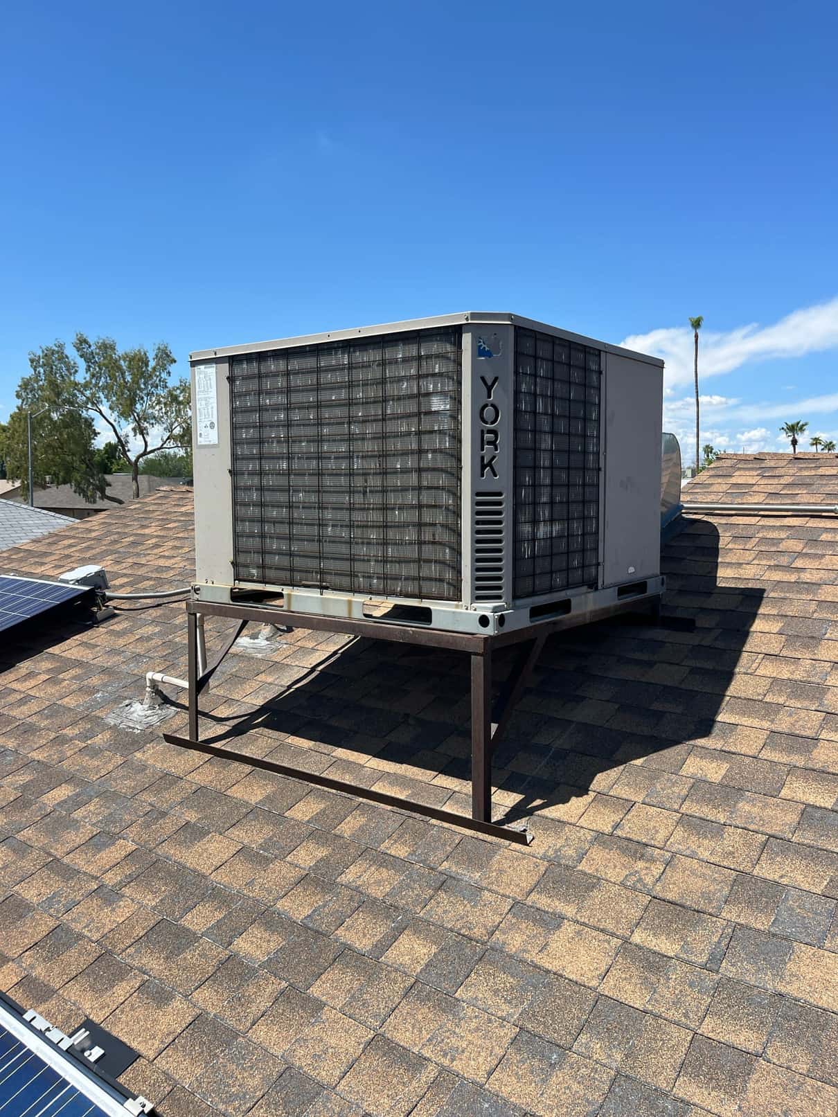 Rooftop packaged air conditioning unit on a metal stand, showing weathered condenser coil fins and rooftop shingles under clear sky.