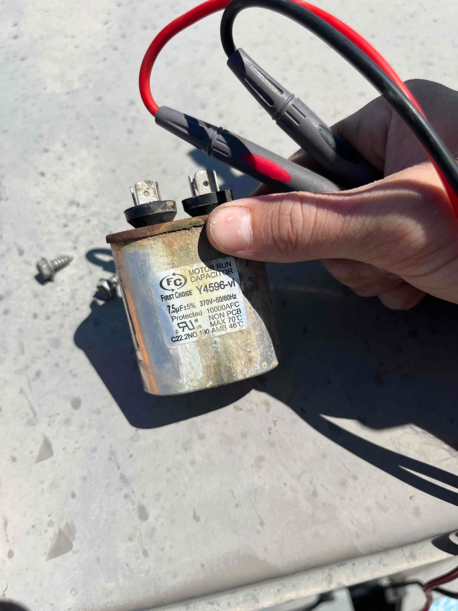 Technician hand holds a removed dual-run capacitor with printed rating 7.5/45 μF and 370/440 VAC beside mounting screws.
