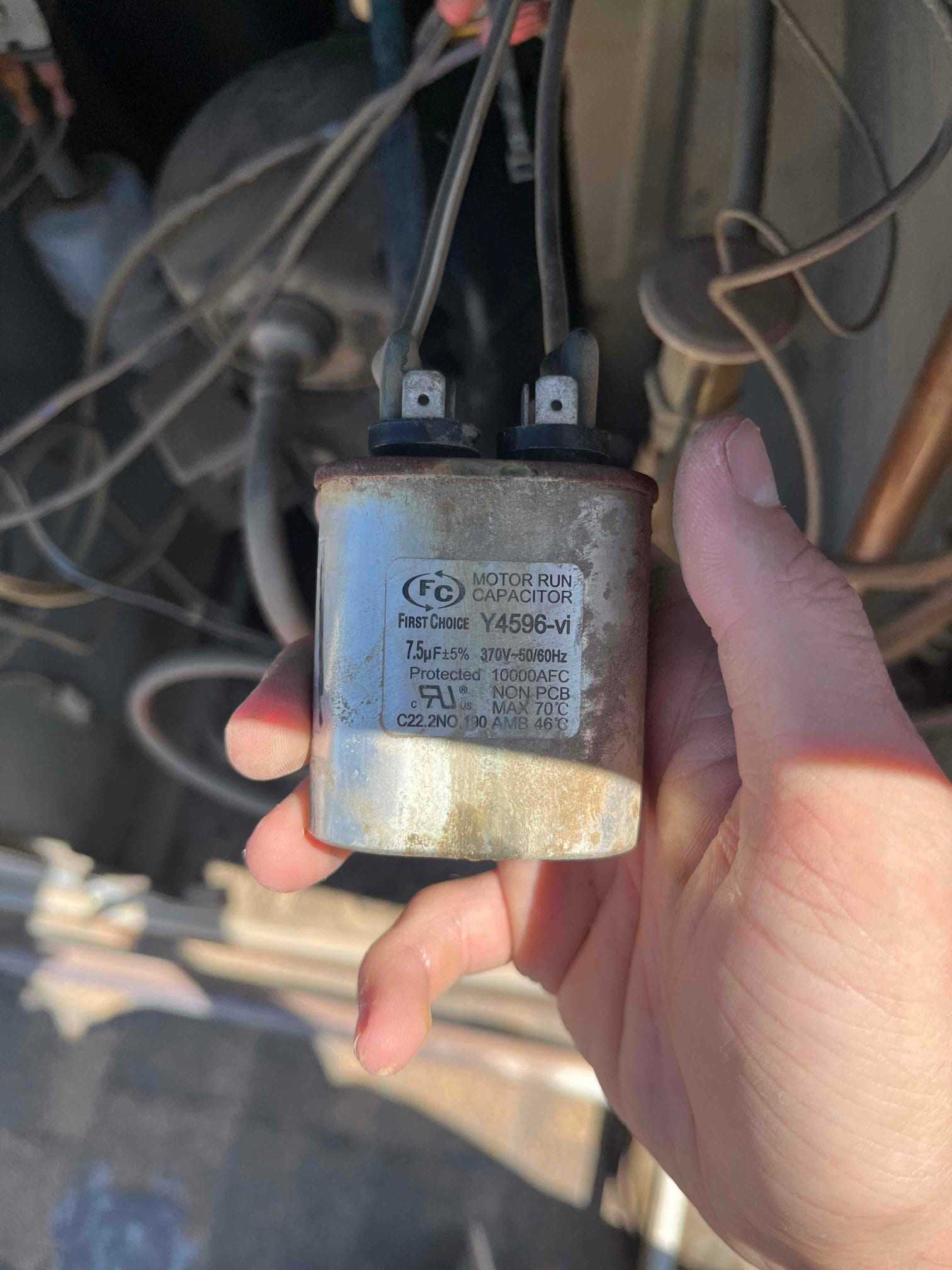 Technician holds a motor run capacitor labeled 7.5 μF ±5%, 370–440 VAC, showing weathered metal can and intact terminals.