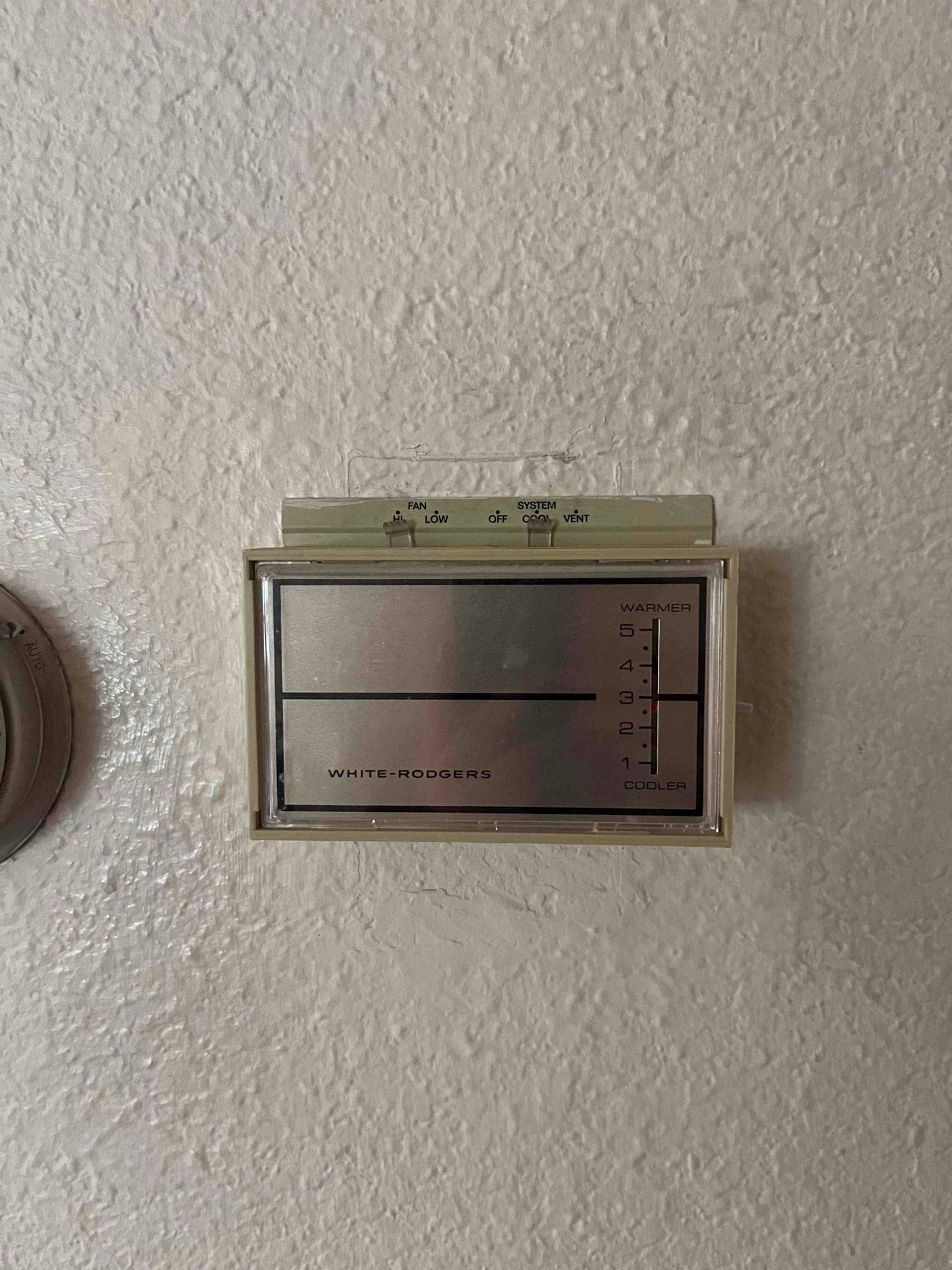 Wall-mounted mechanical thermostat with a sliding temperature lever and visible fan and system mode switches.