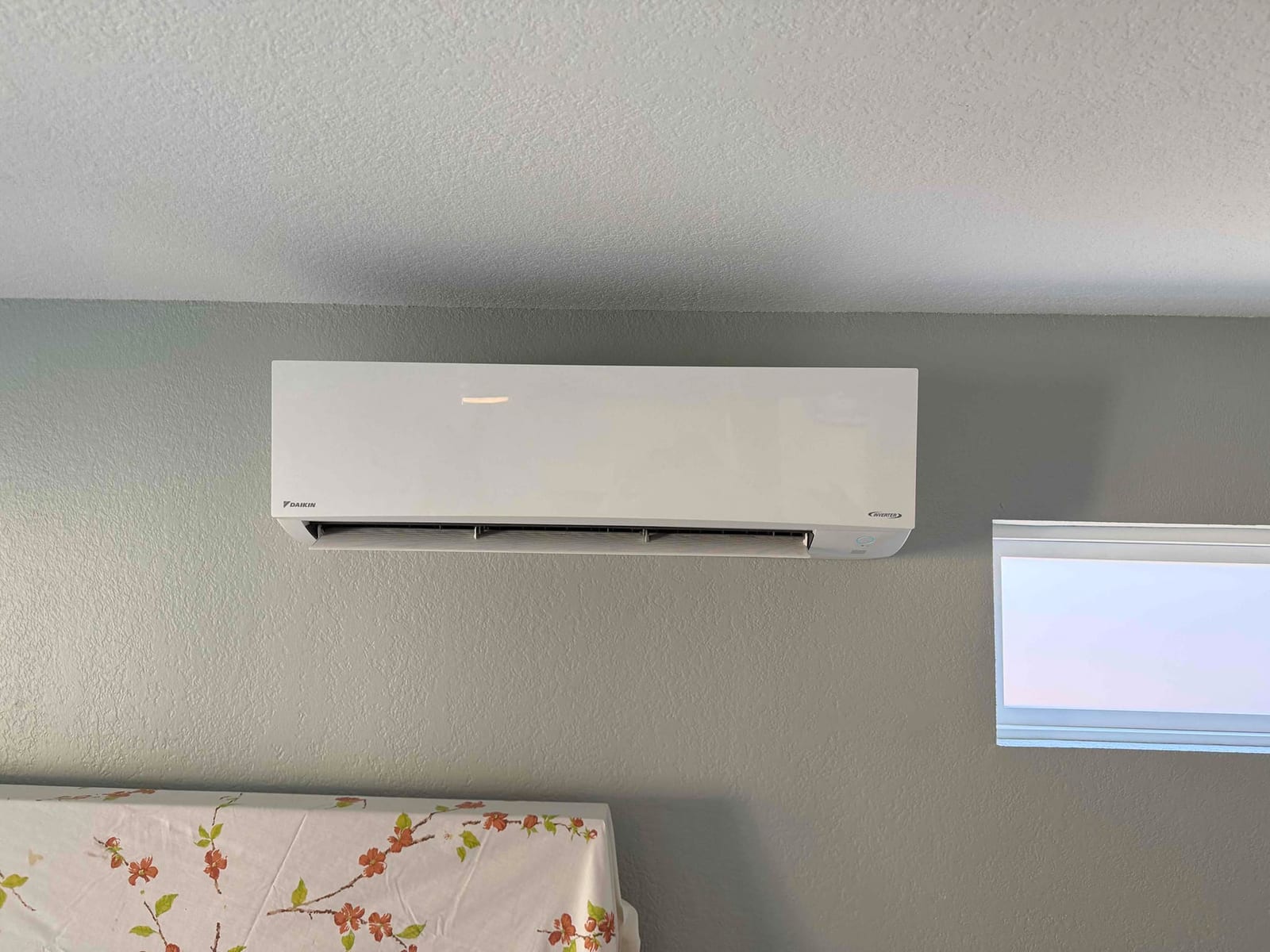 Wall-mounted mini-split indoor unit installed level above a bed, with clean wall penetration concealed and discharge louvers visible.
