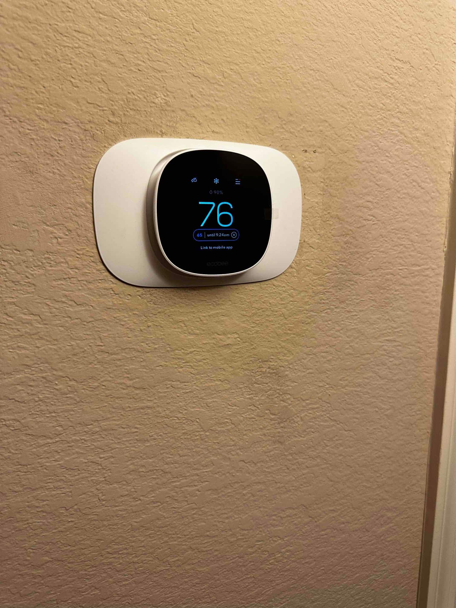 Wall-mounted smart thermostat installed with trim plate, display showing indoor temperature of 76 degrees and active cooling mode icons.