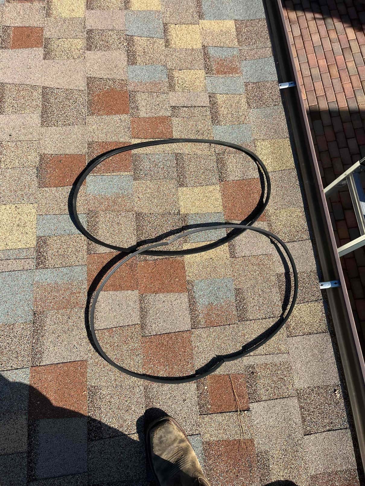 Two worn HVAC drive belts laid on a shingle roof near a gutter, showing cracks and fraying along the edges.