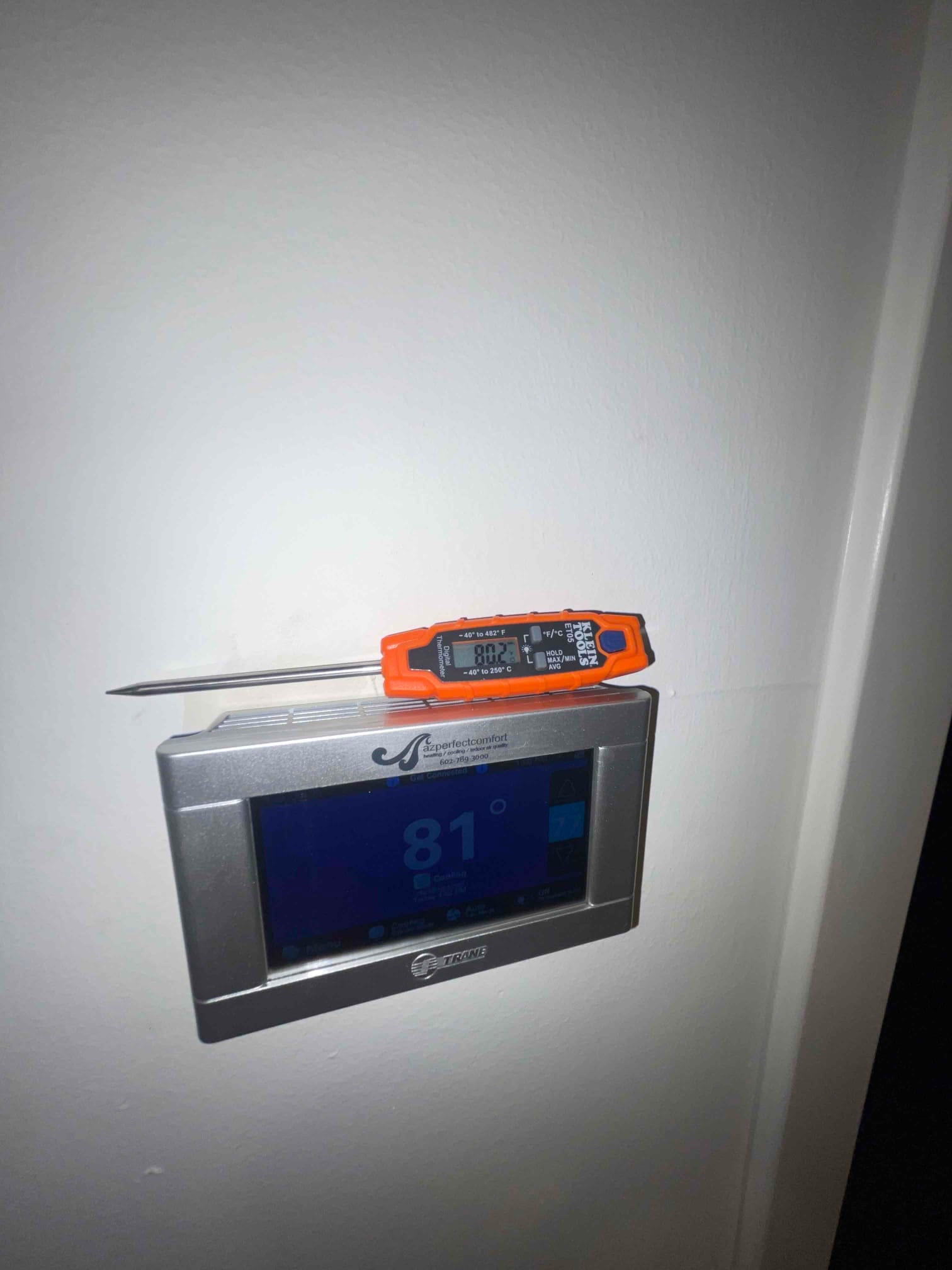 Digital thermometer probe rests on a wall thermostat while measuring air temperature during system checkout.