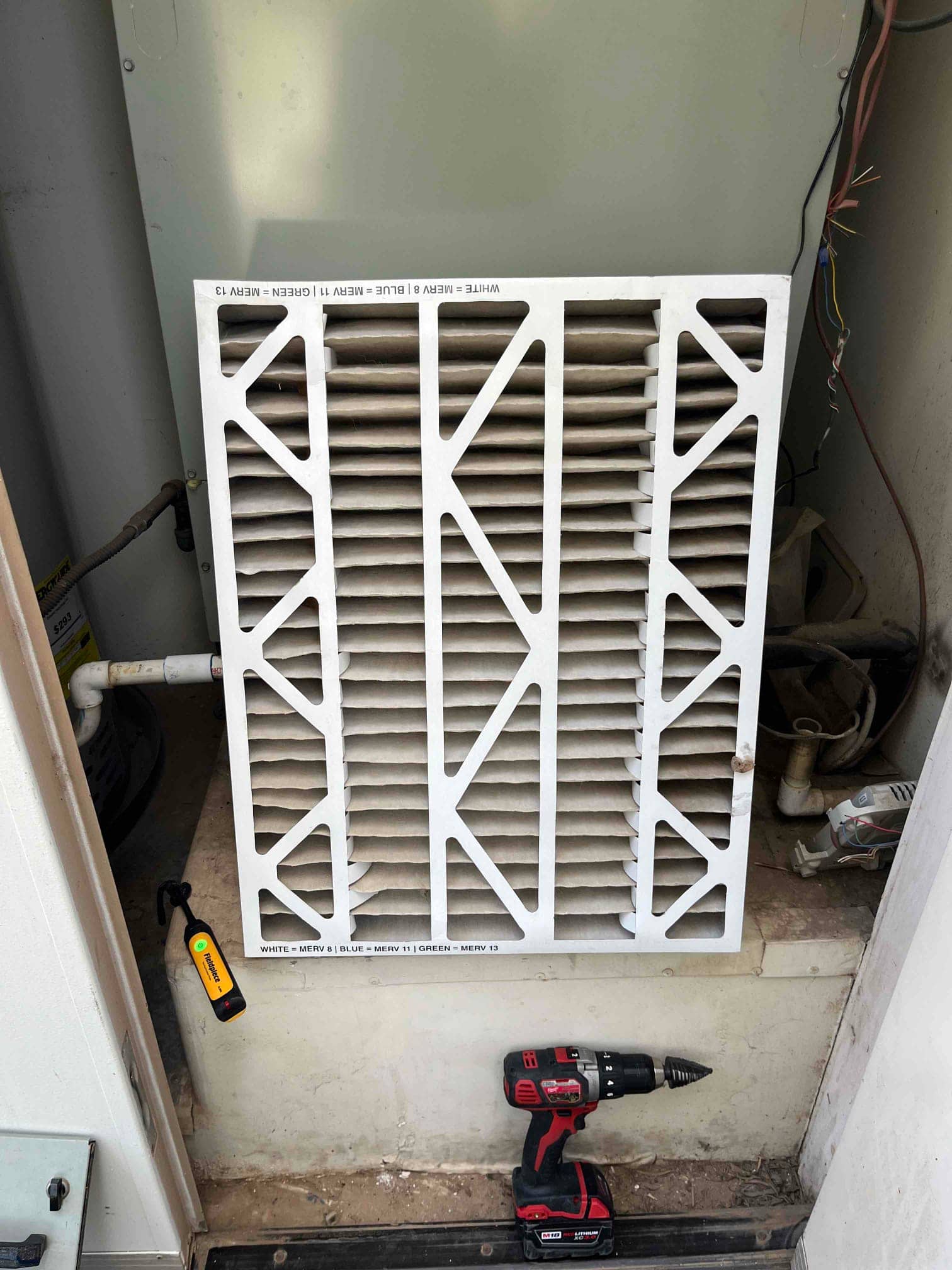 Dirty pleated air filter removed at an indoor air handler compartment, with wiring exposed and tools on the service platform.