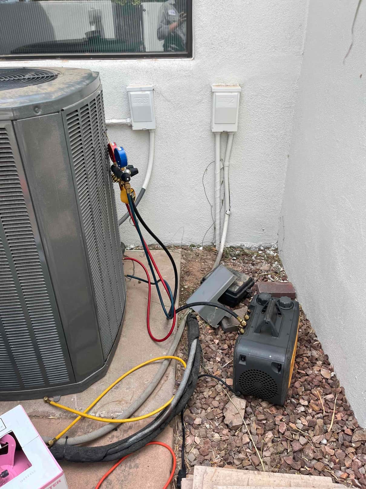 Outdoor condenser with refrigerant manifold hoses connected and a vacuum pump on the ground during service work.