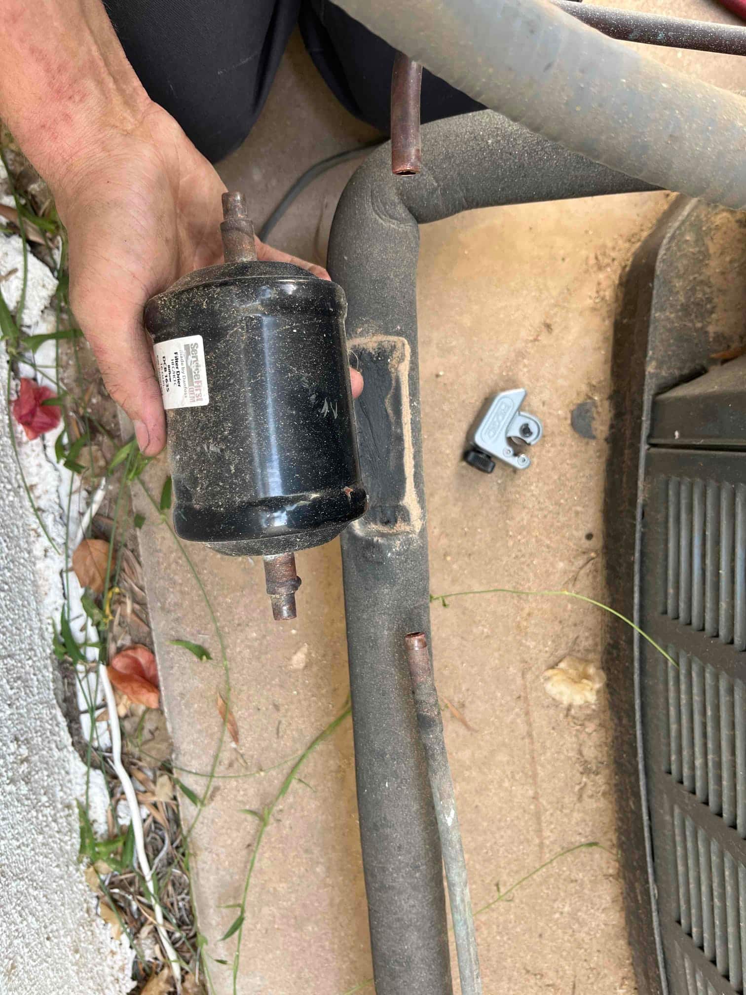 Technician holds a removed inline refrigerant filter-drier next to insulated suction line and outdoor condenser base.
