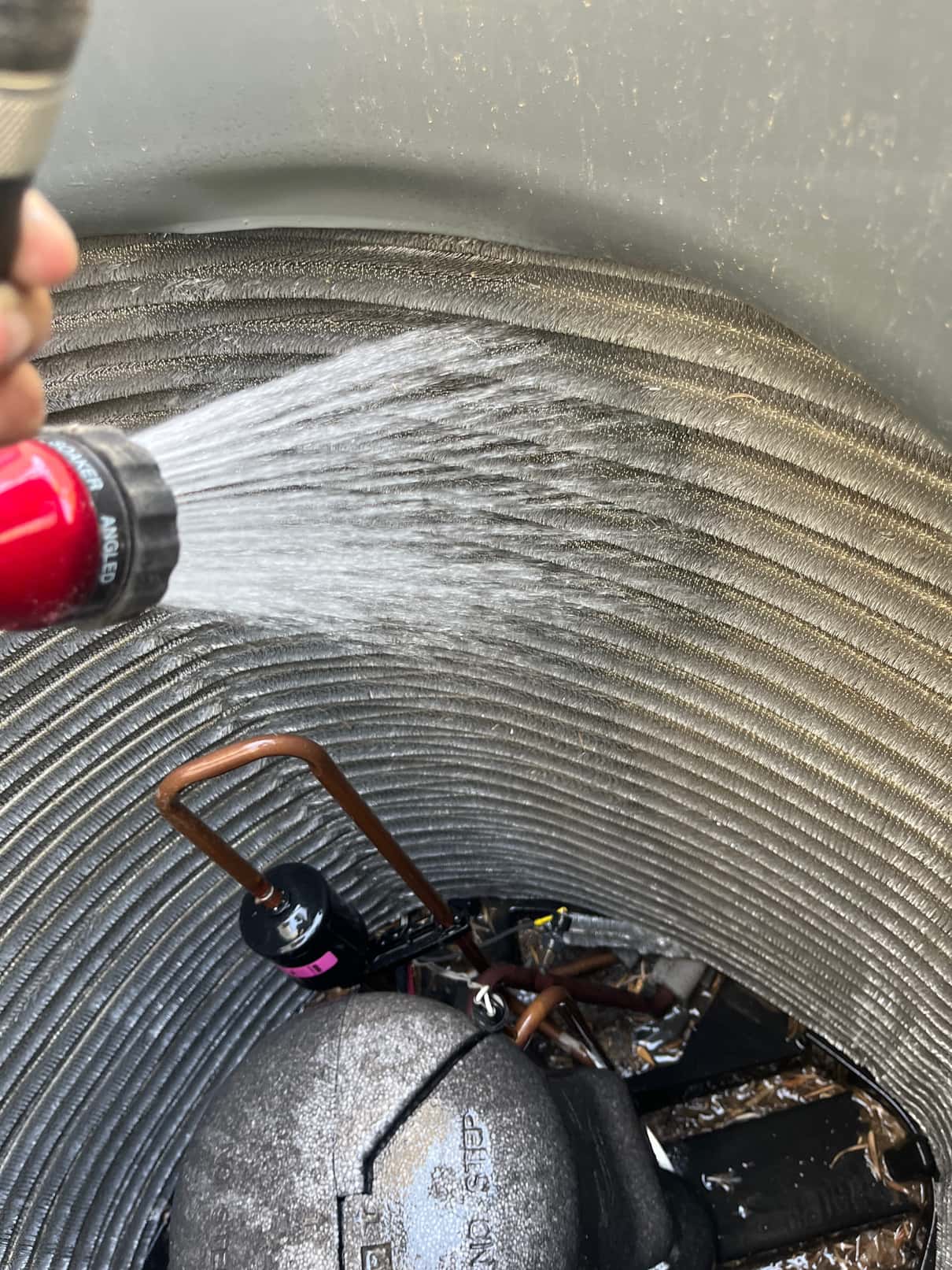 Technician rinses debris from the outdoor condenser coil fins, with compressor top and refrigerant lines visible inside the unit.