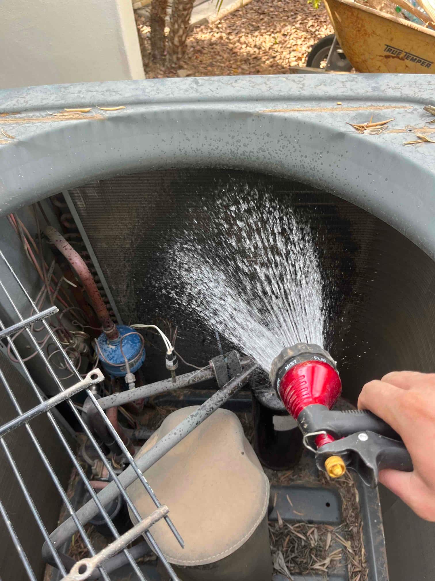 Technician rinses debris from an outdoor condenser coil with a hose, showing water spray across the coil fins and components.