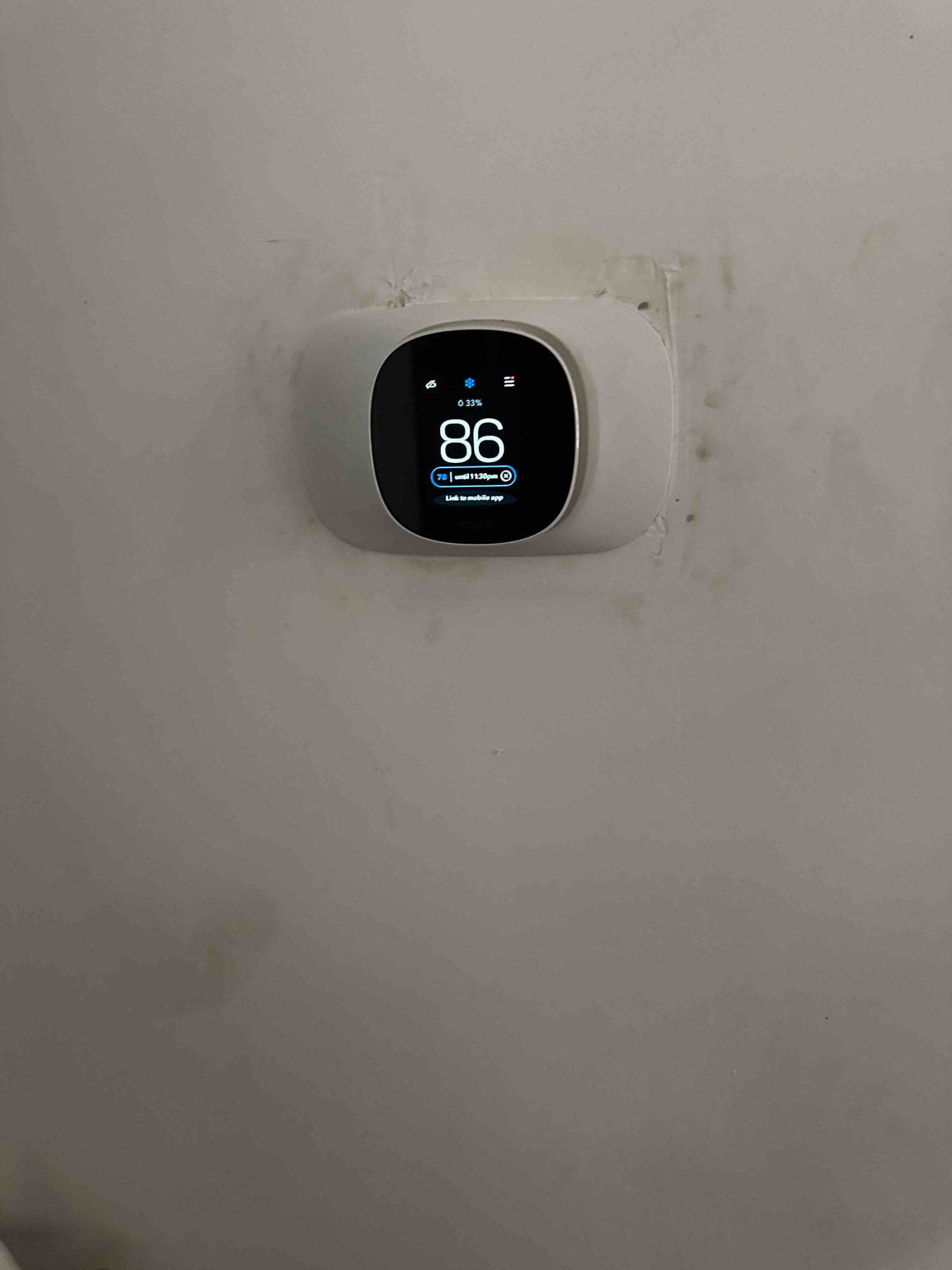 Wall-mounted smart thermostat installed and powered on, displaying indoor temperature and system status on its screen.
