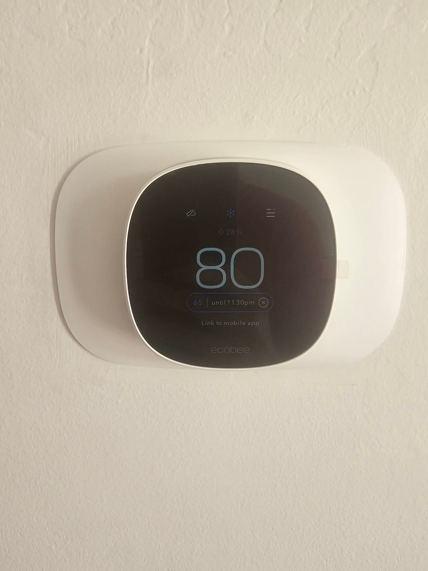 Wall-mounted smart thermostat installed with trim plate, display illuminated and showing current setpoint and system status icons.