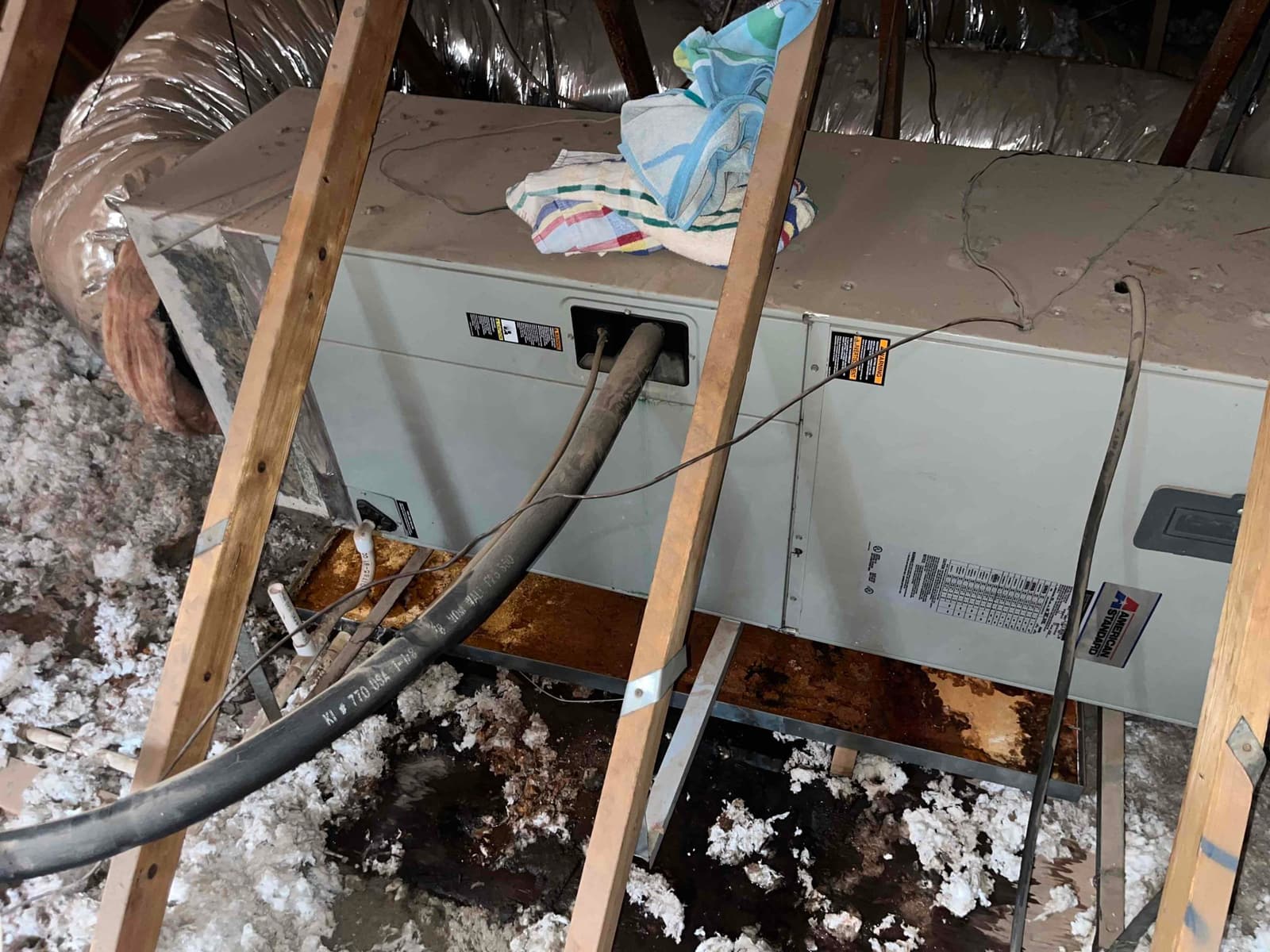 Air handler in attic with removed condensate drain pan, showing water damage and wet insulation around the unit.