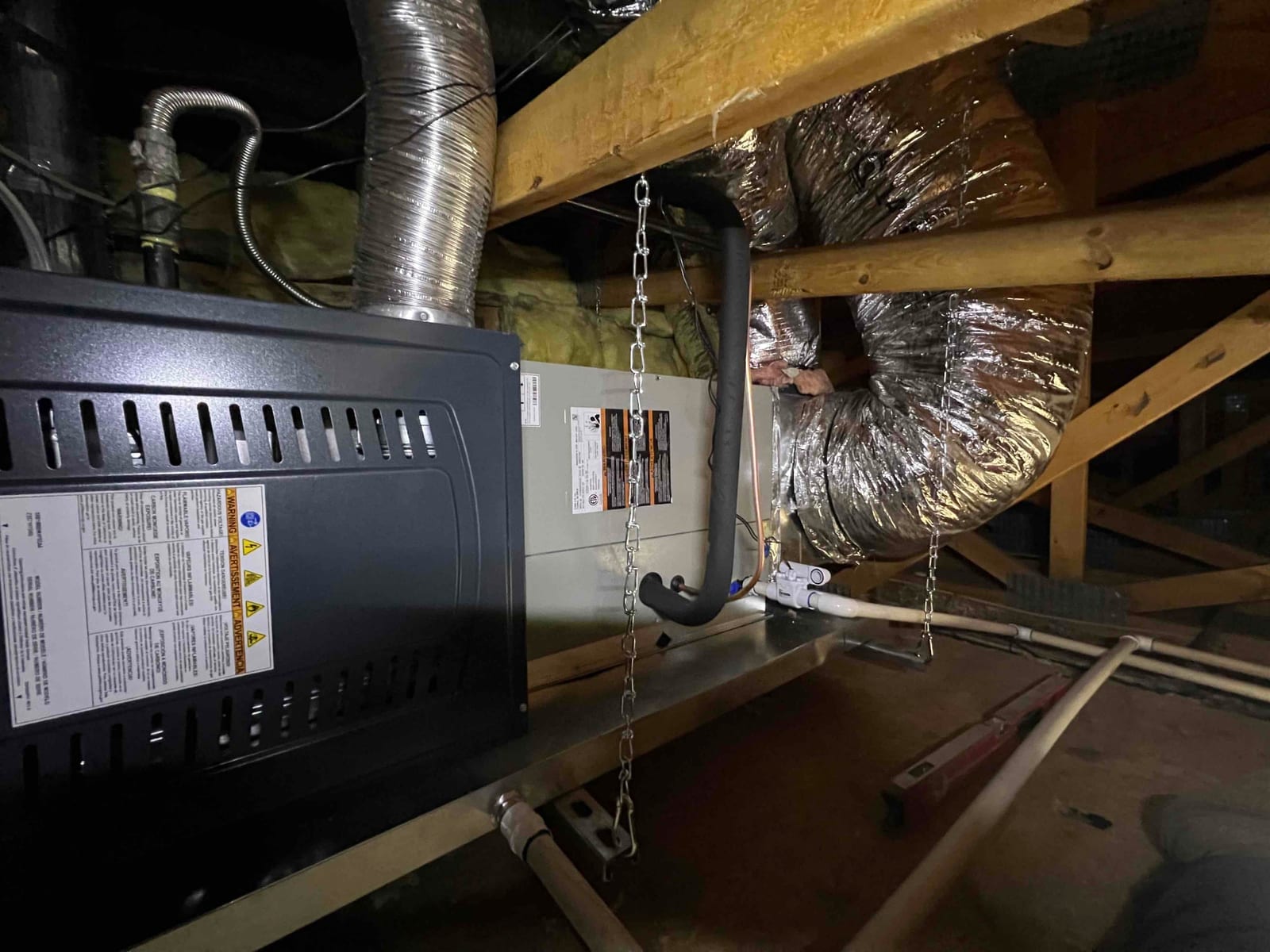 Air handler and furnace installed in attic on a suspended platform with insulated duct connections and condensate drain assembly visible.