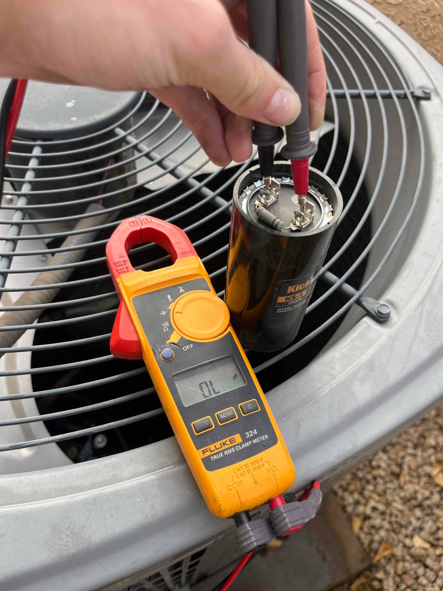 Clamp meter displays 0.1 A while test leads contact terminals on a dual-run capacitor placed on the condenser.