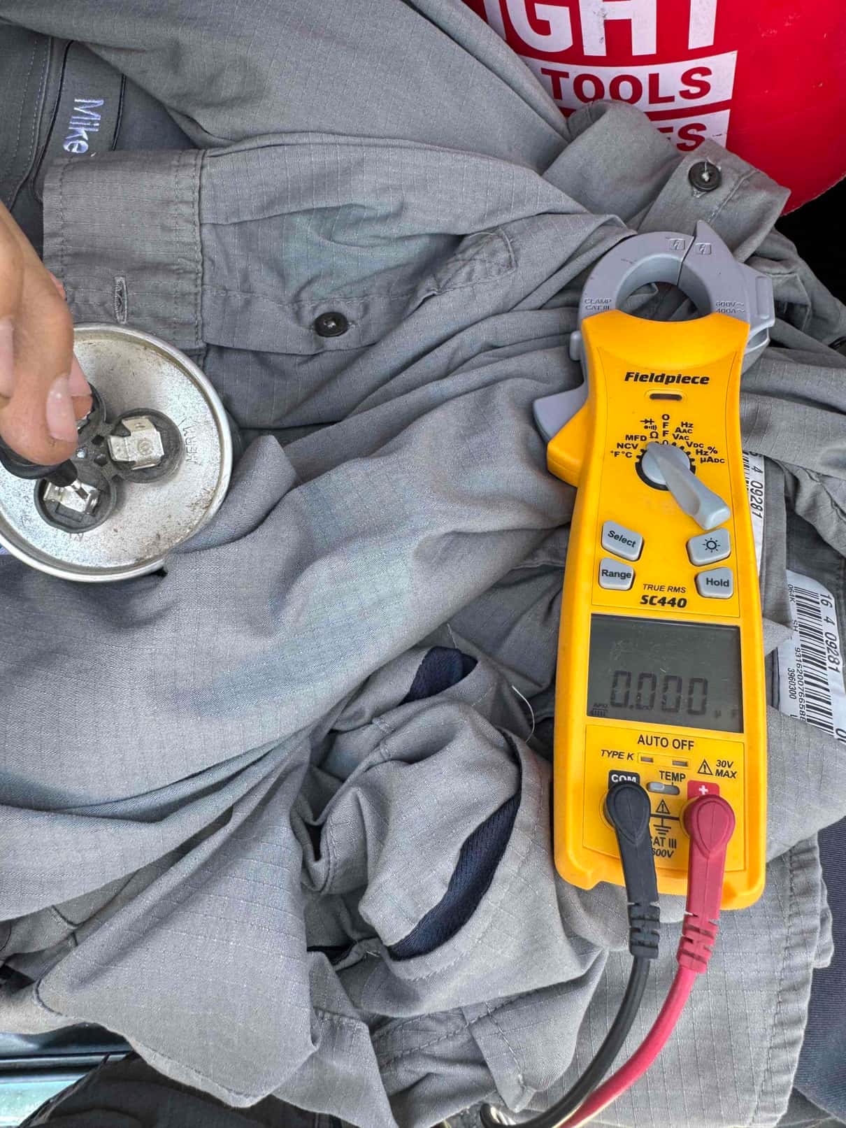 Clamp meter set to AC amps shows 0.000 A while testing alongside a dual-run capacitor with attached wires.