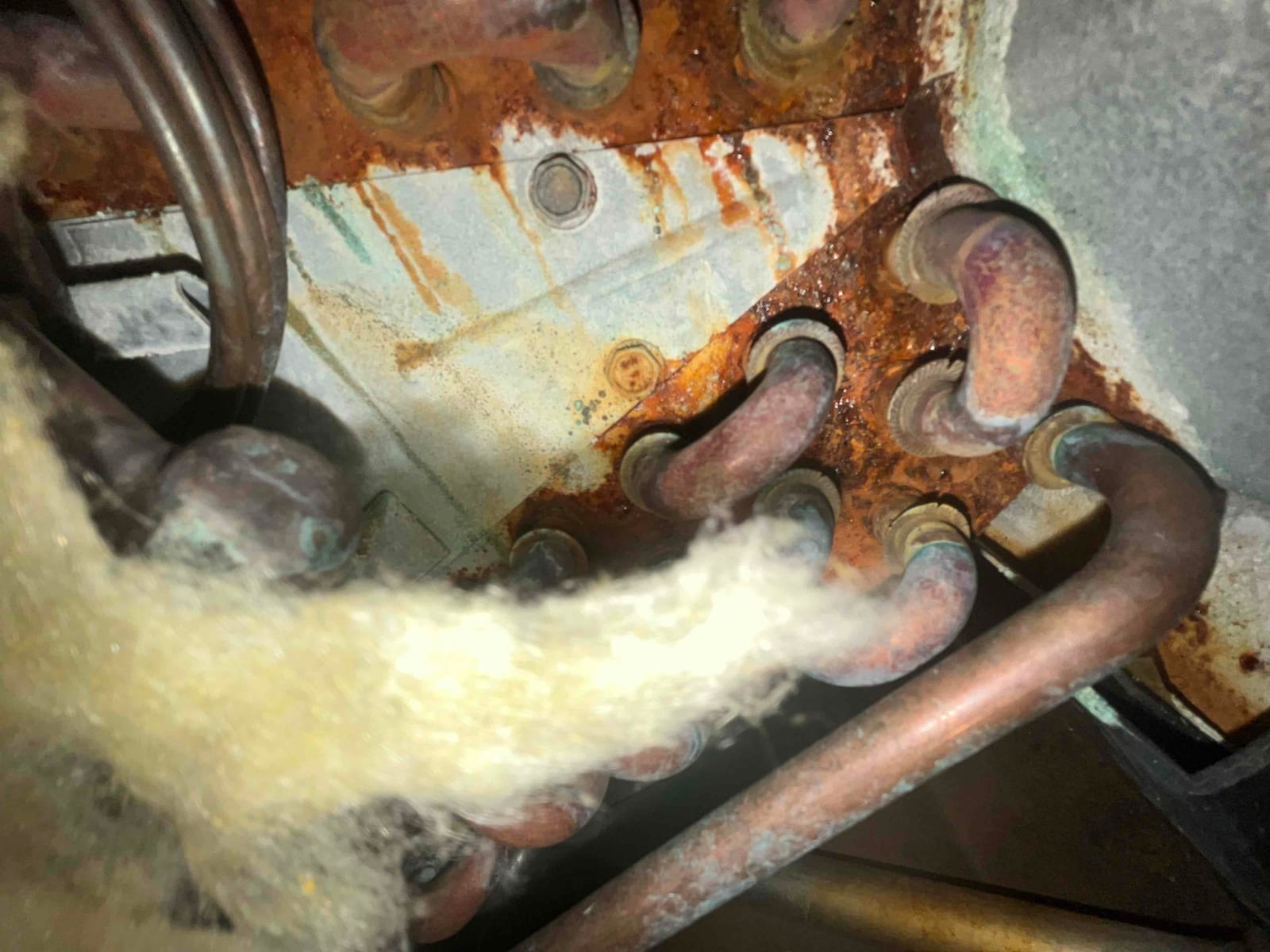 Close-up of corroded refrigerant coil connections with rust staining and insulation fibers near the joint area.